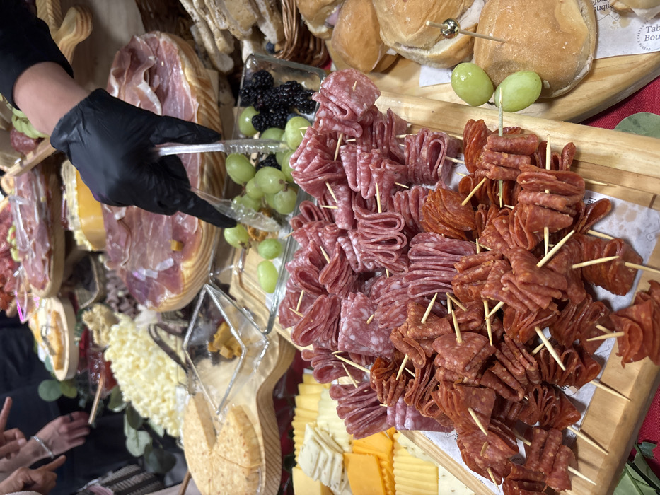 The image displays an elaborate charcuterie and cheese buffet setup, likely situated indoors at an event in Mexico City, Mexico. In the immediate foreground, a wooden serving board lined with white paper holds numerous individual skewers, each containing multiple folded slices of cured meats: a lighter pinkish-red variety and a darker reddish-orange variety. To the right, a second wooden board presents small bread rolls, one with a decorative pick piercing two green grapes. Adjacent to this, a gloved hand, clad in a black nitrile glove and part of a black long-sleeved garment, uses clear plastic tongs to serve green grapes from a glass dish that also contains blackberries.
Further into the mid-ground, a selection of sliced cheeses, including yellow and white blocks, is arranged on a tray, alongside a round, textured cheese on a separate wooden board. Multiple large wooden boards in the background are laden with fanned slices of cured ham, possibly prosciutto or jamón. Other visible food items include a pile of shredded white cheese, a bowl of red jam, and a basket of various bread items. Parts of two additional individuals are discernible in the upper left background, with one hand adorned with a silver bracelet and gesturing towards the food display. Green leafy garnishes are interspersed among the dishes, which are set upon a red surface.