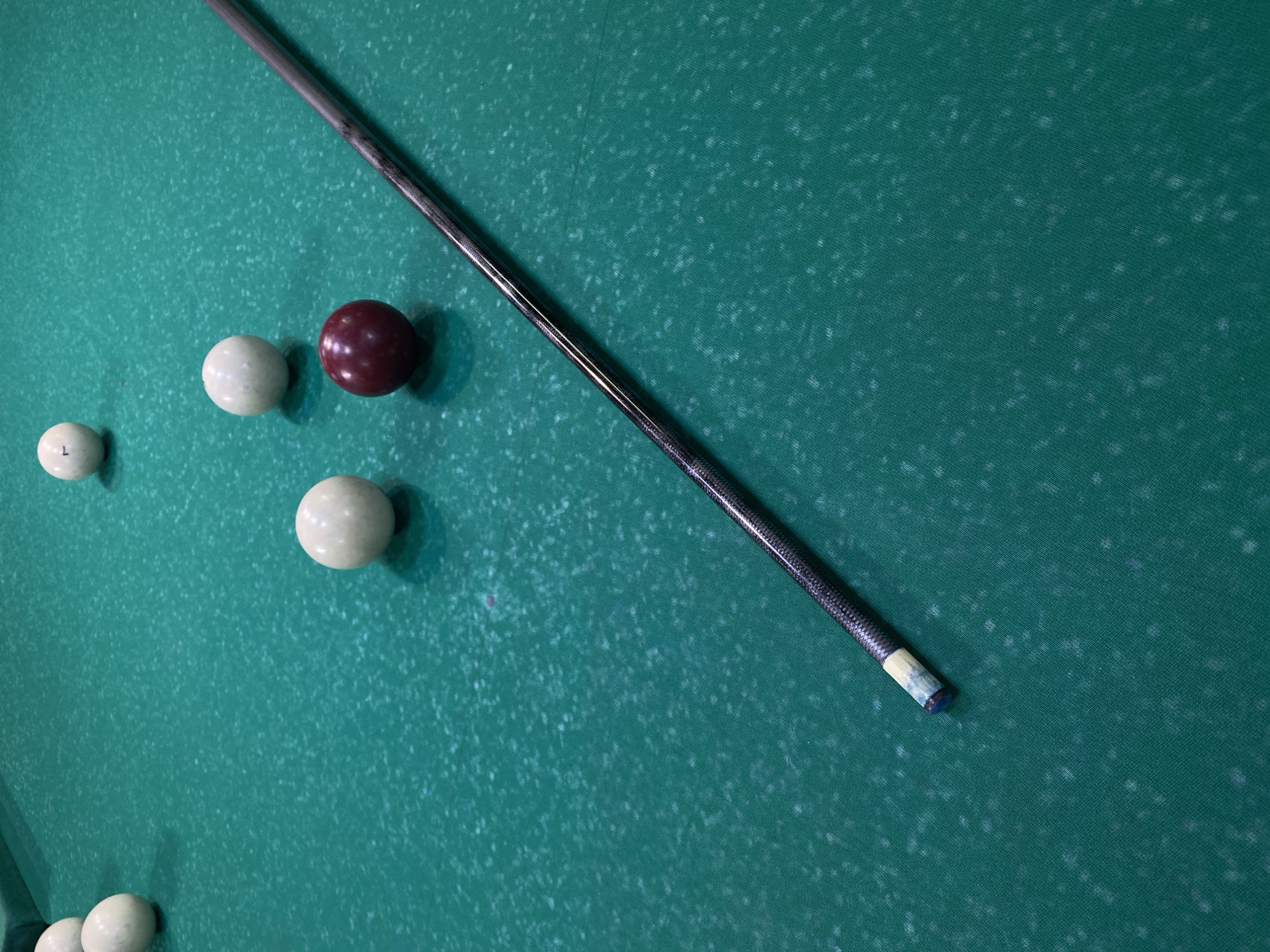 The image depicts a green billiard table. There are several balls scattered on the table: three white balls and one maroon ball. A billiard cue rests diagonally across the table, with the tip near the upper right corner of the image. One of the white balls has a letter "L" on its surface. The table's green cloth exhibits a textured pattern. The location, Chaikovsky, Russia is not discernible in the image.