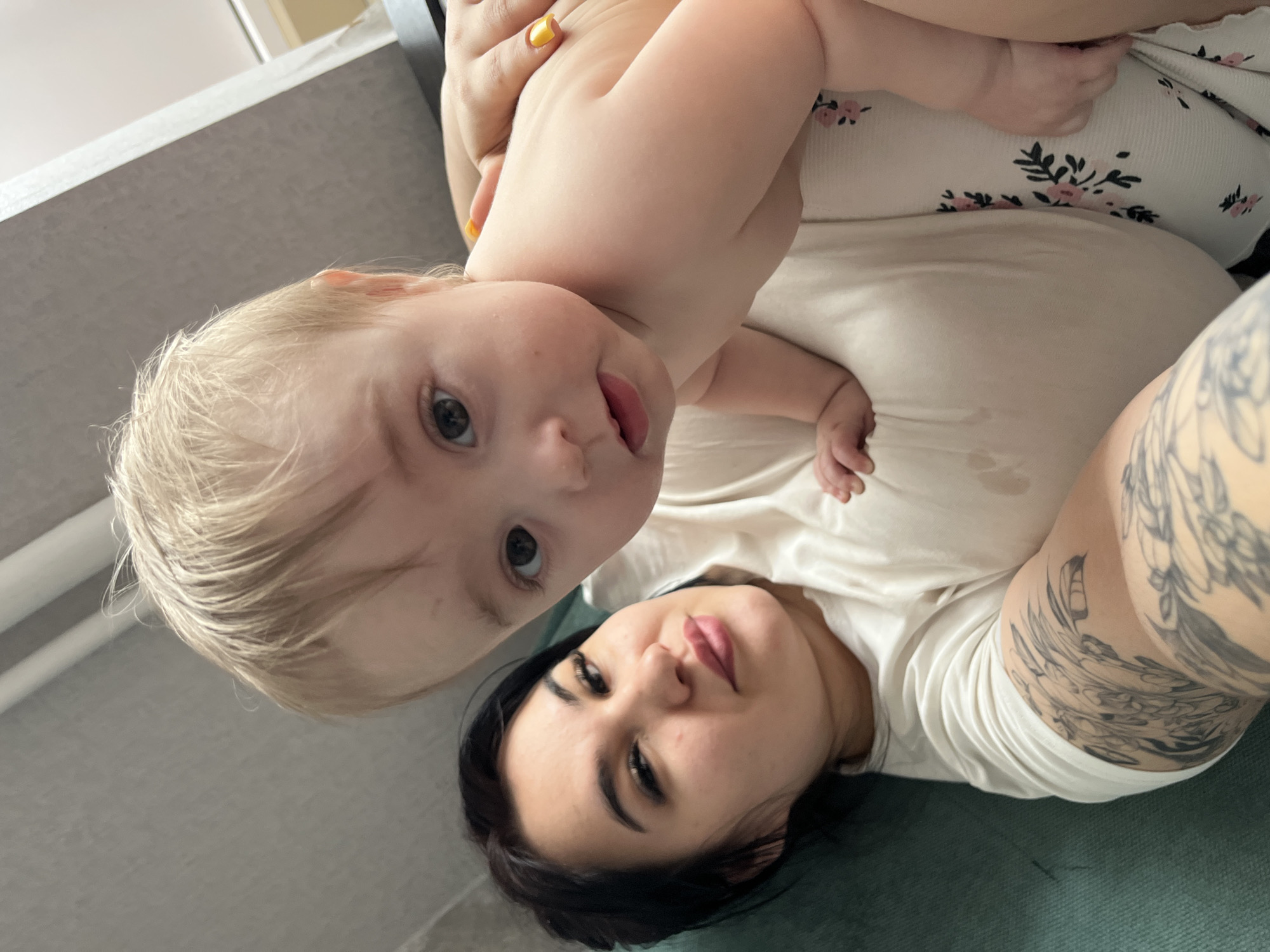 A woman with dark hair and a tattoo on her arm is lying down with a baby. The baby has blonde hair and is sticking its tongue out. The location is likely in a home. The city of Chaikovsky, Russia cannot be confirmed.