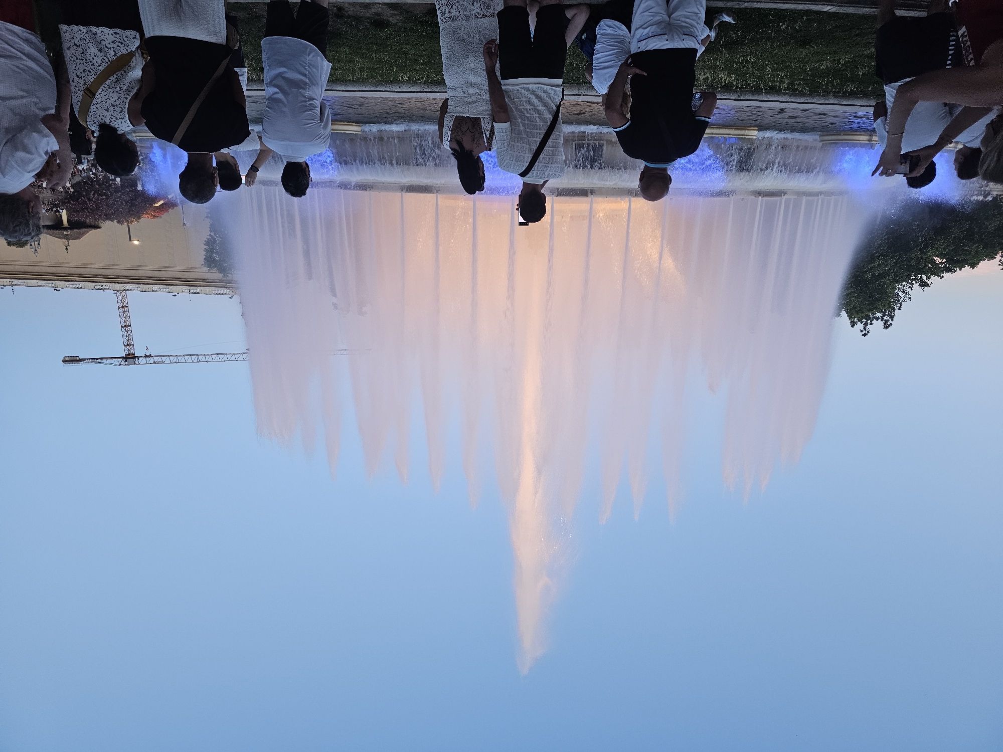 The image depicts the Magic Fountain of Montjuïc in Barcelona, Spain, during dusk. A large, illuminated fountain is the central focus, with numerous water jets reaching upwards and a prominent central jet. Blue and white lights illuminate the water, and the sky is a clear, pale blue. A crowd of people stands in the foreground, facing the fountain and observing it. Many are taking photographs with their mobile phones. The people are dressed in casual summer attire. In the background, behind the fountain and the crowd, a large building and a construction crane are visible. Trees and greenery flank the sides of the fountain. The overall atmosphere appears to be one of public enjoyment and spectacle.