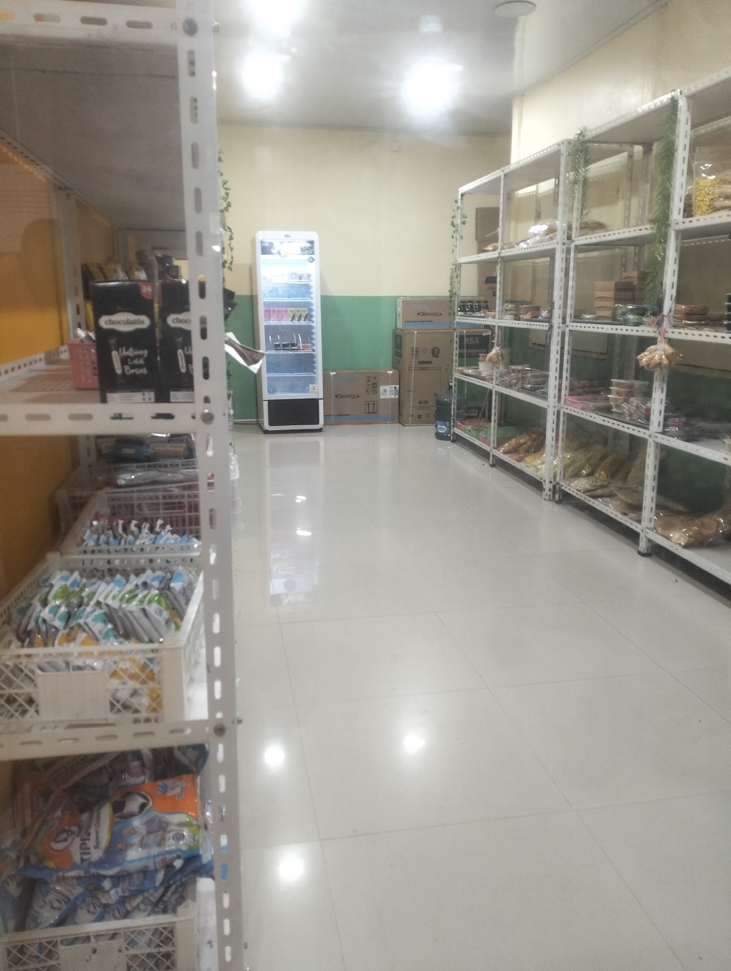The image shows the interior of a small grocery store in Air Jamban, Indonesia. The store is lined with white metal shelves filled with various food products. On the left, there are shelves stocked with packaged snacks and drinks, including a large black box labeled "Chocolatos." In the center, a white beverage cooler is visible. To the right, more shelves display packaged goods, some in clear plastic bags. Cardboard boxes are stacked near the shelves. The floor is tiled and reflective, with lighting fixtures visible on the ceiling. The overall impression is of a modest, well-stocked convenience store.