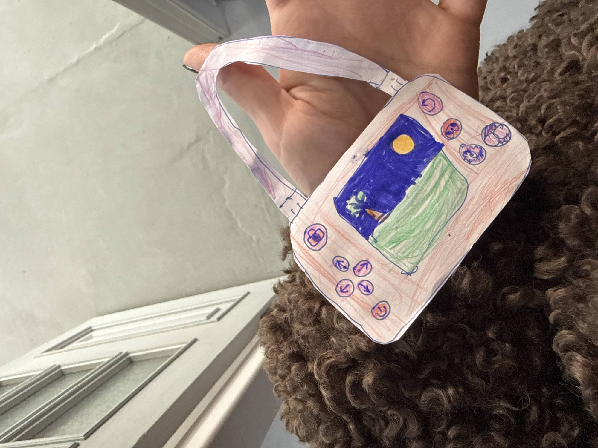 A person's hand holds up a child's paper craft, designed to resemble either a miniature handbag or a vintage handheld gaming device. The craft features a crudely drawn and colored "screen" area with a blue sky, a yellow sun, and green ground or landscape. Surrounding this screen are several circular 'buttons' with various simple designs, including arrows and spirals, drawn in blue, purple, and orange. The paper handle of the craft is also visible, made from a strip of paper. The hand holding the craft has manicured nails, possibly with dark polish.
A large, dense mass of curly, brown, and dark brown fur or fabric occupies the right and lower portions of the image, suggesting the presence of a fluffy pet or a textured coat. In the background, to the left, is a light-colored, possibly off-white, wall or ceiling. Towards the bottom left, part of a white door or window frame with decorative molding and glass panes is visible. The scene appears to be an indoor setting in Krasnoyarsk, Russia. No visible text is present.