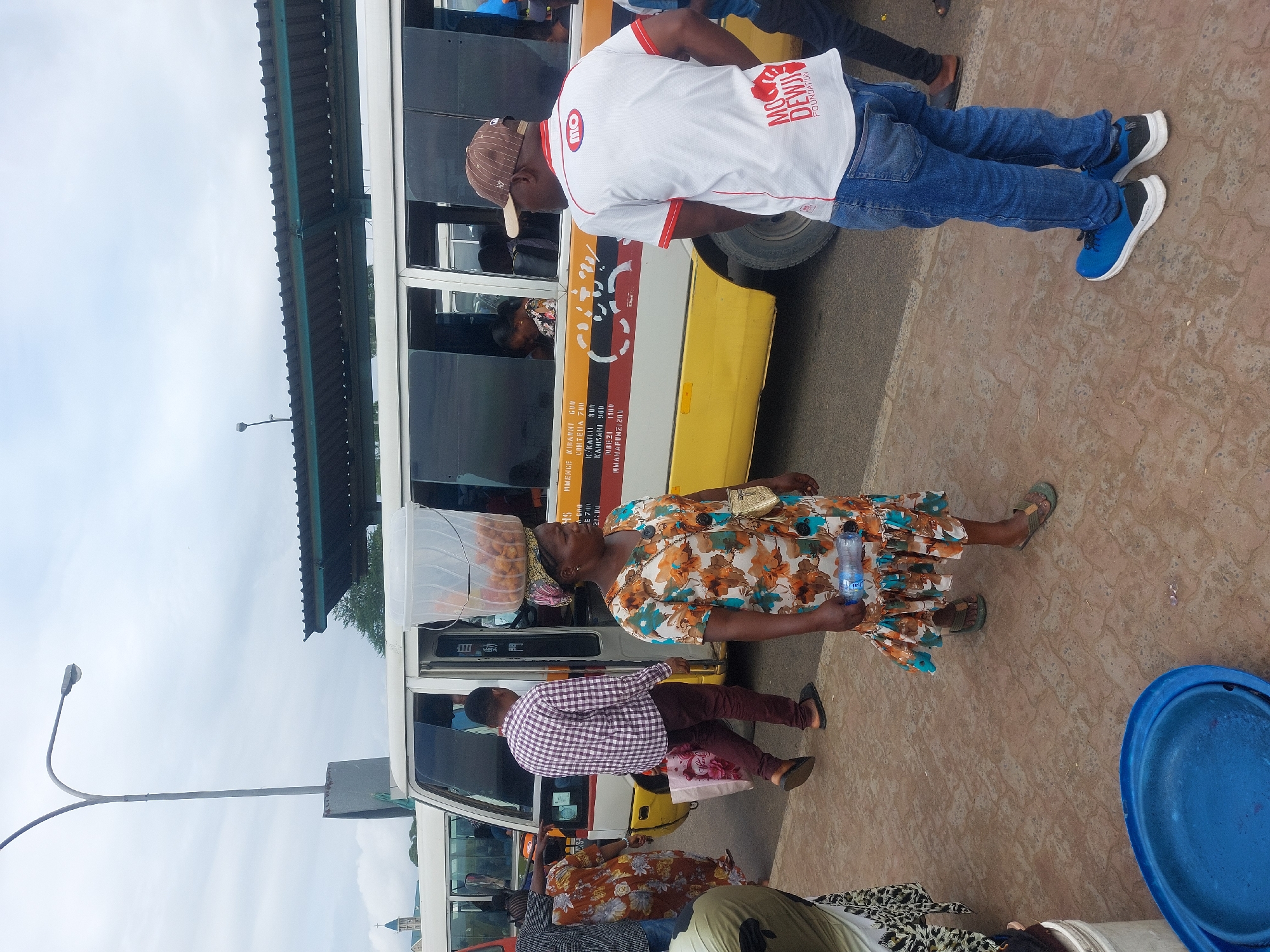 The image captures a bustling street scene in Dar es Salaam, Tanzania, evidenced by the crowded minibus and the Swahili text on its side indicating various local destinations such as Mwenge Kibarani, Conteina, K/Kariakoo, Kahi Sahi, and Mbezi. Pedestrians surround the bus, with a woman balancing a container of what appears to be doughnuts or fried dough on her head. She also holds a water bottle in her hand.
A man in jeans and a white t-shirt with the inscription "MO DEWJI FOUNDATION" is standing near the front of the bus. Other individuals are seen waiting or walking around the bus, including one in a checkered shirt. There is a blue plastic basin on the ground near the edge of the frame. The backdrop shows a partly cloudy sky, a streetlight, and parts of the building. The ground appears to be paved with patterned tiles.