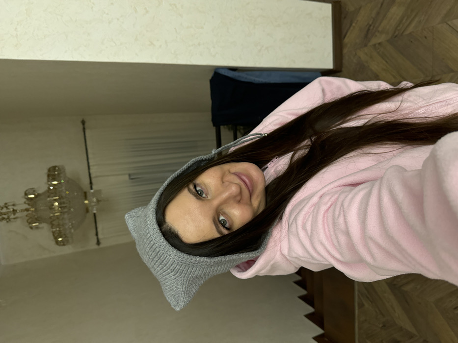 The image portrays a female individual in an indoor residential setting within Vysokinichi, Russia. The woman, appearing to be in her 20s or 30s with long dark brown hair and blue-green eyes, is positioned slightly off-center and looks directly at the camera with a slight smile. She is wearing a light grey knit hat with prominent cat ears and a light pink, possibly fleece, hooded zip-up jacket.
The background includes a high ceiling from which a large, ornate, gold-toned chandelier with multiple light sources is suspended. Light-colored, textured wallpaper covers the walls. To the left, a wooden staircase with visible treads ascends. Behind the subject's head, a window is visible, covered by light-colored sheer curtains and an upper dark curtain rod. The floor exhibits dark wood parquet patterns. In the far-right background, a dark object, possibly an item of clothing or furniture, is visible against the wall.