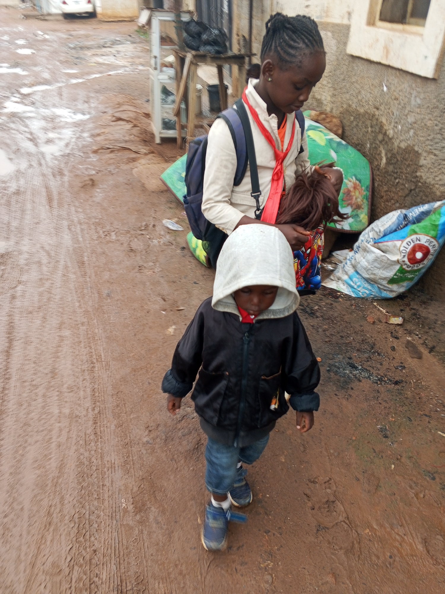 The image depicts a young child, wearing a hooded black jacket, blue jeans, and sneakers, walking on a wet, muddy dirt road. The child is looking downwards. Behind the child, an older child, possibly a girl with braided hair, is seated on a colorful mat, holding a doll. She is wearing a light-colored jacket over an orange shirt and a red lanyard. A blue backpack is on her shoulders. To the right, a large, partially filled blue and white plastic bag with the words "GOLDEN PENNY FOODS" is visible. The environment appears to be an outdoor setting, likely a residential or informal commercial area in Abuja, Nigeria, given the muddy conditions and the style of the structures and objects present. The ground shows tire tracks, suggesting vehicle traffic.