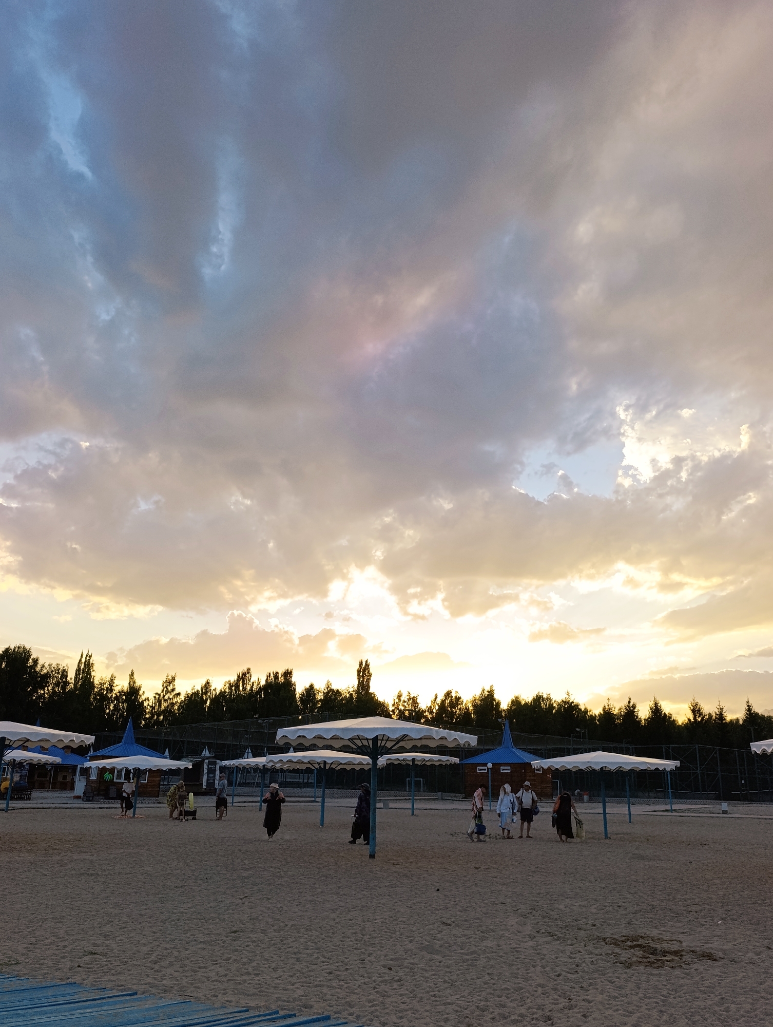 The image depicts a sandy beach area, possibly part of a recreation facility, in Bayet, Kyrgyzstan, under a cloudy sky with sunset hues. Several white beach umbrellas with blue accents are erected on the sand. In the background, a line of dark green trees forms a dense forest. A few small buildings with blue roofs are visible among the trees.
Several people are present on the beach. On the left, a group of three individuals, one possibly a child, are standing near a building. Further to the right, a group of four people are walking together. A single individual in a dark outfit is walking towards the right side of the frame. On the far right, another individual is seen walking away from the camera. The sand is lightly textured, with some areas appearing slightly darker, suggesting dampness or variations in the sand itself. The overall scene suggests a relatively quiet end to the day, with the last visitors enjoying the beach as the sun sets.