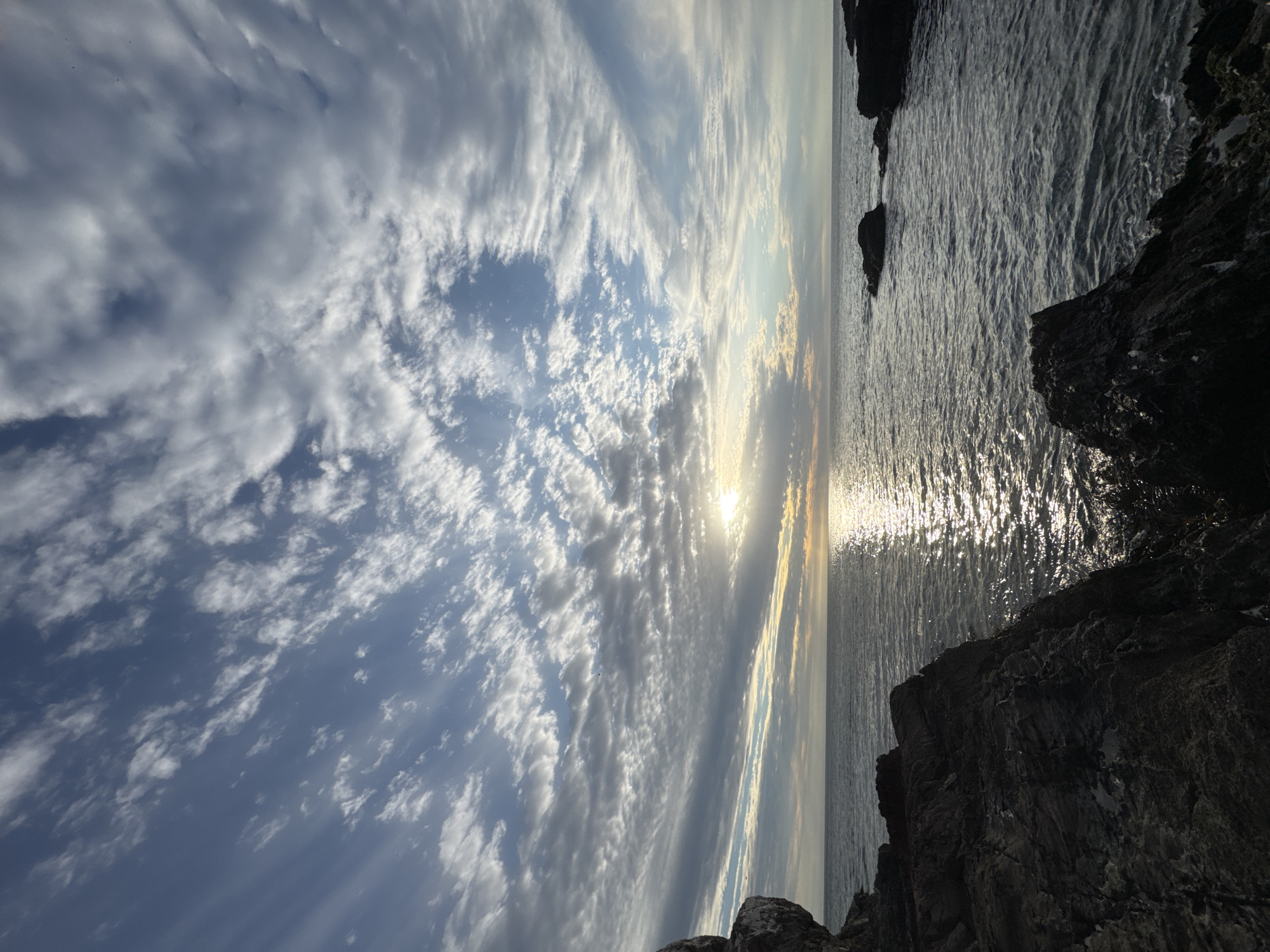The image depicts a seaside scene with a dramatic sky and rocky foreground. The sky is filled with a mix of clouds, ranging from dense and dark to wispy and light, with a clear blue patch visible in between. The sun is partially obscured by the clouds, creating a bright, shimmering effect on the water's surface. The water itself is dark and choppy, with visible reflections of the sunlight. In the foreground, large, dark rocks jut out from the edge of the water, their texture rough and jagged. The image is oriented sideways, with the sky and clouds on the left and the water and rocks on the right. The location is unspecified and unknown.