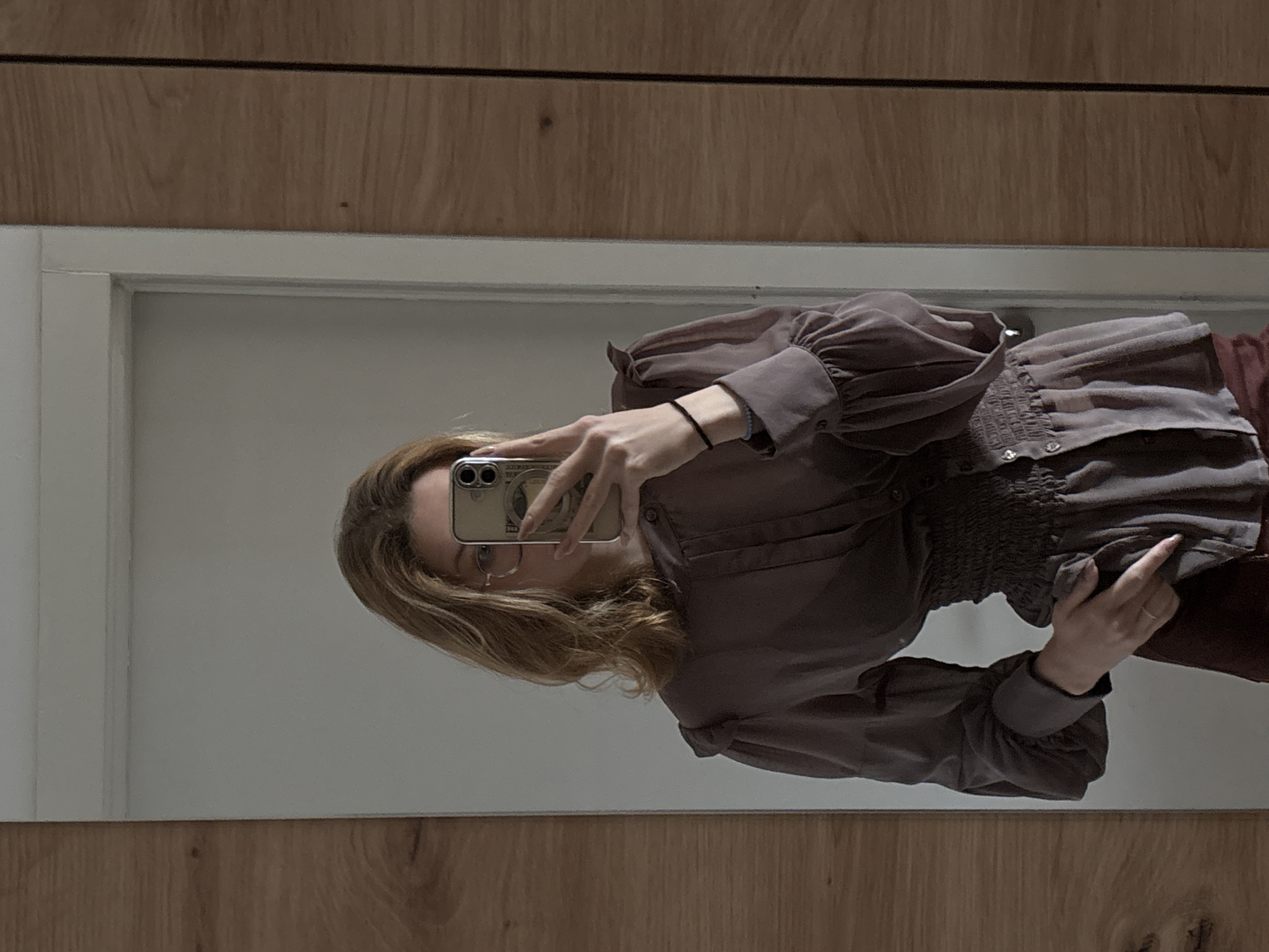 A mirror reflects a woman in a purple blouse and maroon pants. She holds a phone up to take a picture of herself in the mirror. The setting is indoors, with light wood paneling on the wall around the mirror. There is a white frame for the mirror. The location is unknown.