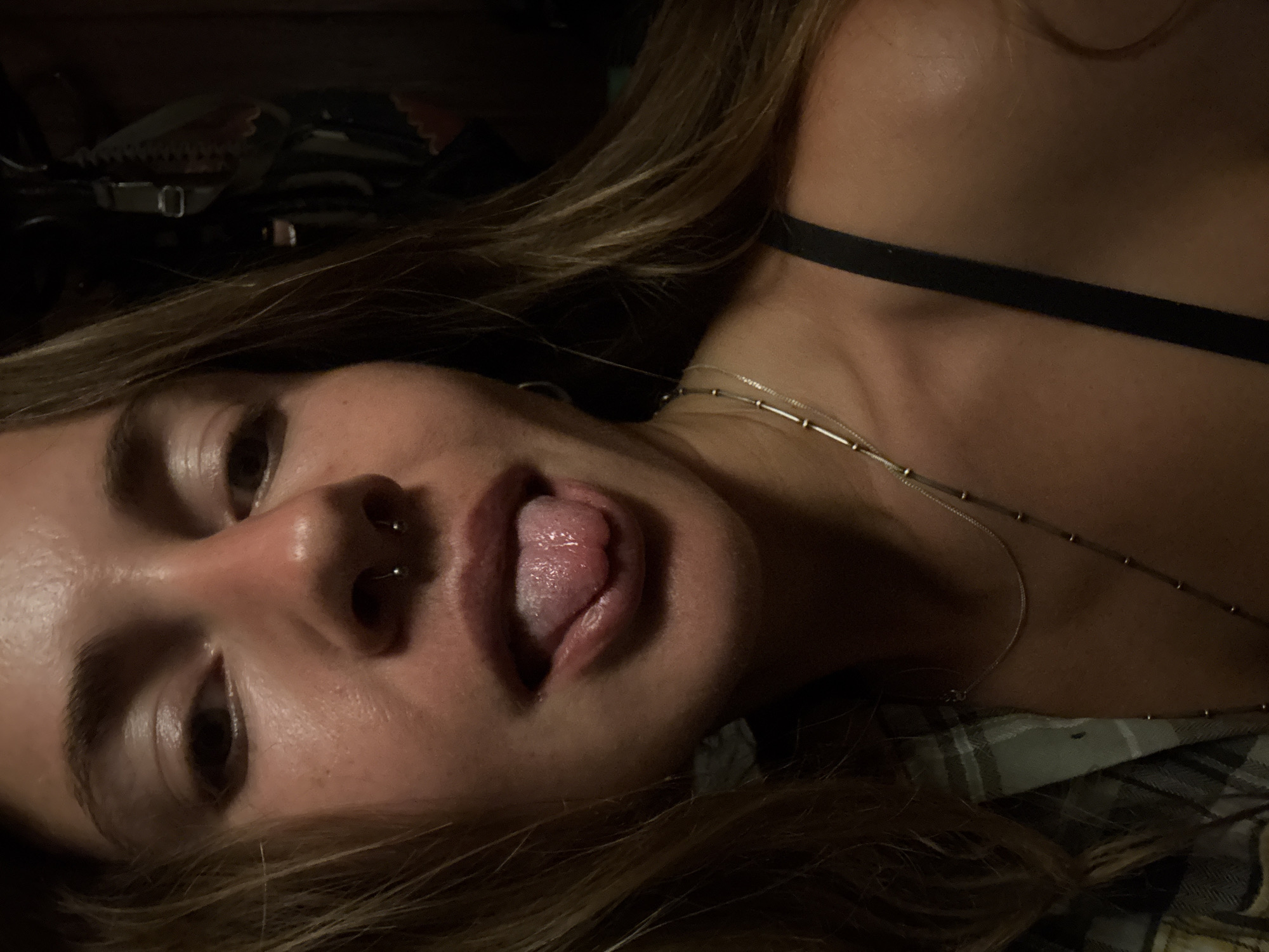 The image is a close-up, selfie-style shot of a young woman with her tongue sticking out. She has light brown hair that is parted to her left and falls around her face. She is wearing a silver septum ring. Her skin appears to have some texture and shine, possibly due to sweat or makeup. She is wearing a black bra strap visible on her right side and a plaid shirt on her left. Two silver necklaces are visible around her neck. The background is dark and appears to be indoors, with some indistinct objects and textures suggesting a cluttered or storage area. The lighting is dim, with the primary light source appearing to be from the front, illuminating her face.
