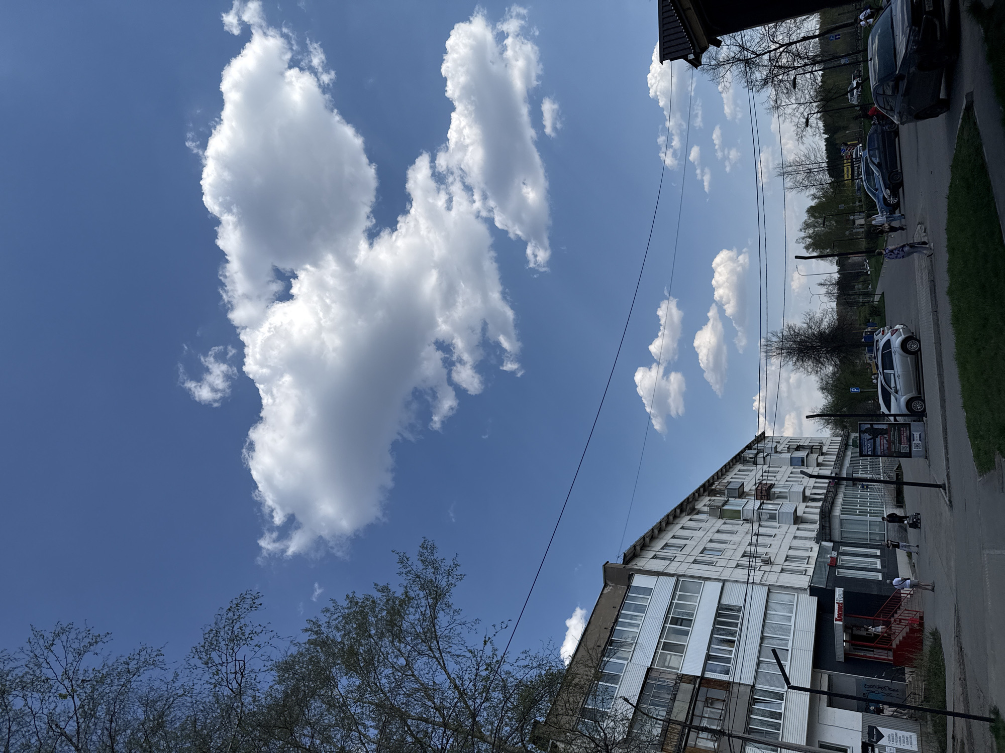 The image depicts a street scene in an unidentified city. A tall apartment building with white balconies dominates the right side of the frame. A few people are walking on the sidewalk adjacent to the building. A street with parked cars runs parallel to the building. Power lines stretch across the sky. Above the building, a bright blue sky is dotted with cumulus clouds. Bare tree branches are visible in the foreground.