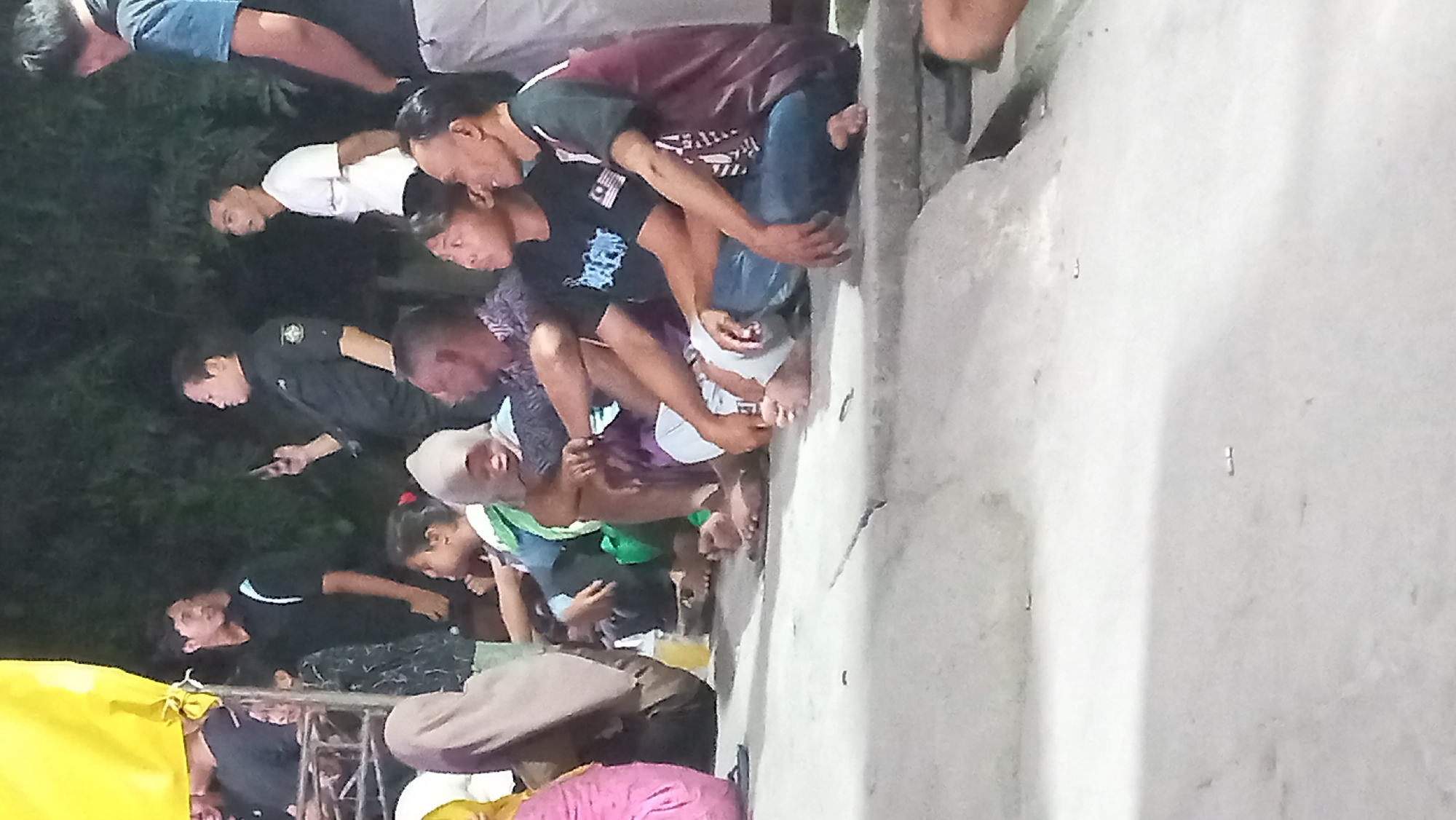 A group of people are sitting on a concrete surface outdoors at night. The people are mostly young adults and appear to be of Southeast Asian descent. Some are wearing casual clothing, while others are in darker, uniform-like attire. One person in the background is holding a smartphone. The environment appears to be a public gathering or event, possibly in a rural or semi-urban setting in Kedungmegarih, Indonesia, given the presence of lush vegetation in the background and the style of dress. The lighting suggests artificial illumination from an unseen source, casting shadows on the concrete.