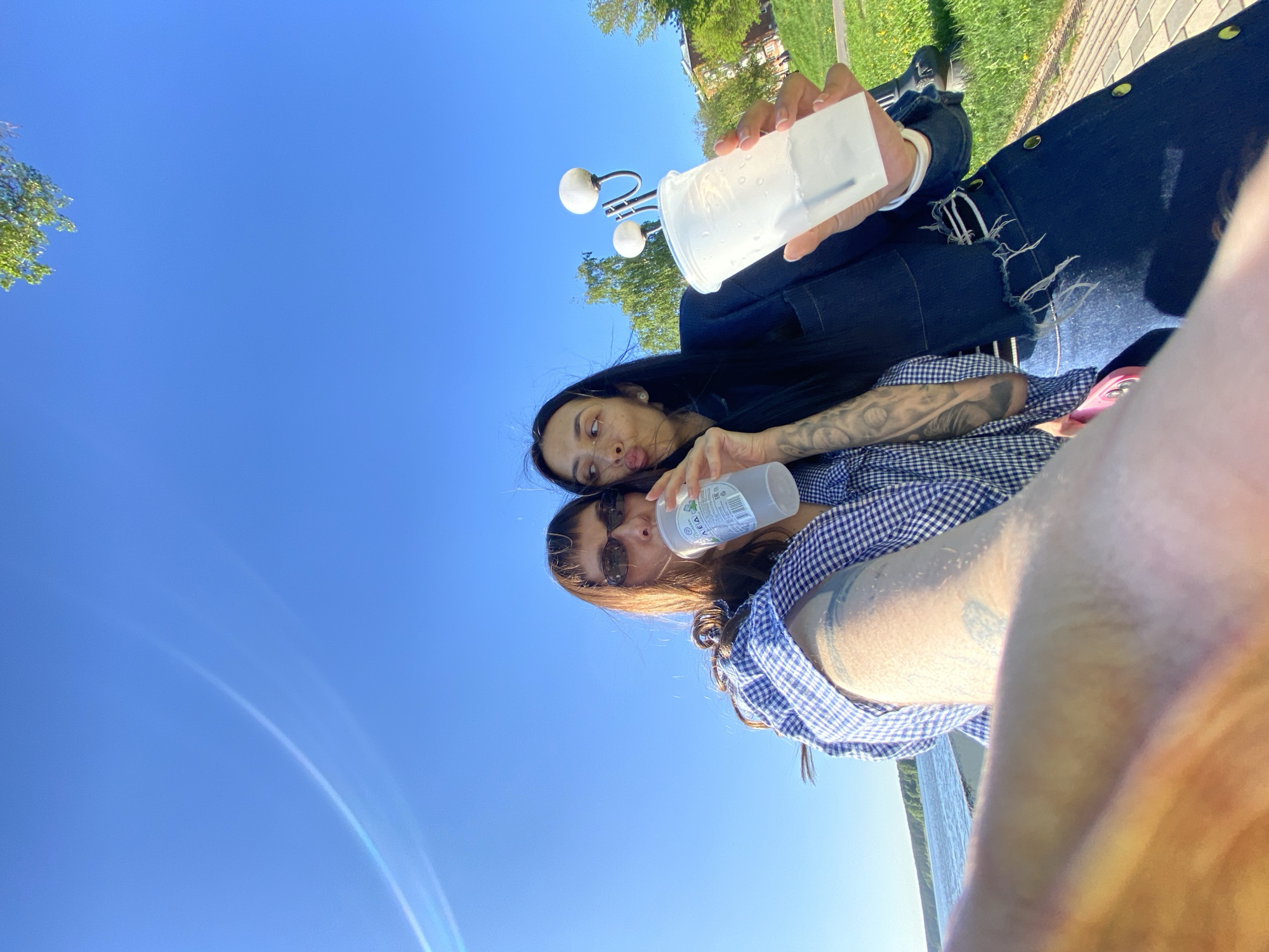 Two individuals are seated outdoors on what appears to be a bench. The view is angled upwards towards the sky. The person closer to the camera wears sunglasses and a blue and white checkered shirt. A hand, presumably belonging to this person, holds a translucent container. The other person is behind them, wearing a dark-colored garment, and also holding a translucent container. Tattoos are visible on their arm.
The environment includes a clear blue sky, trees, and possibly a light fixture in the background. Based on the visual information, the precise location within Votkinsk, Russia, cannot be determined with certainty.
