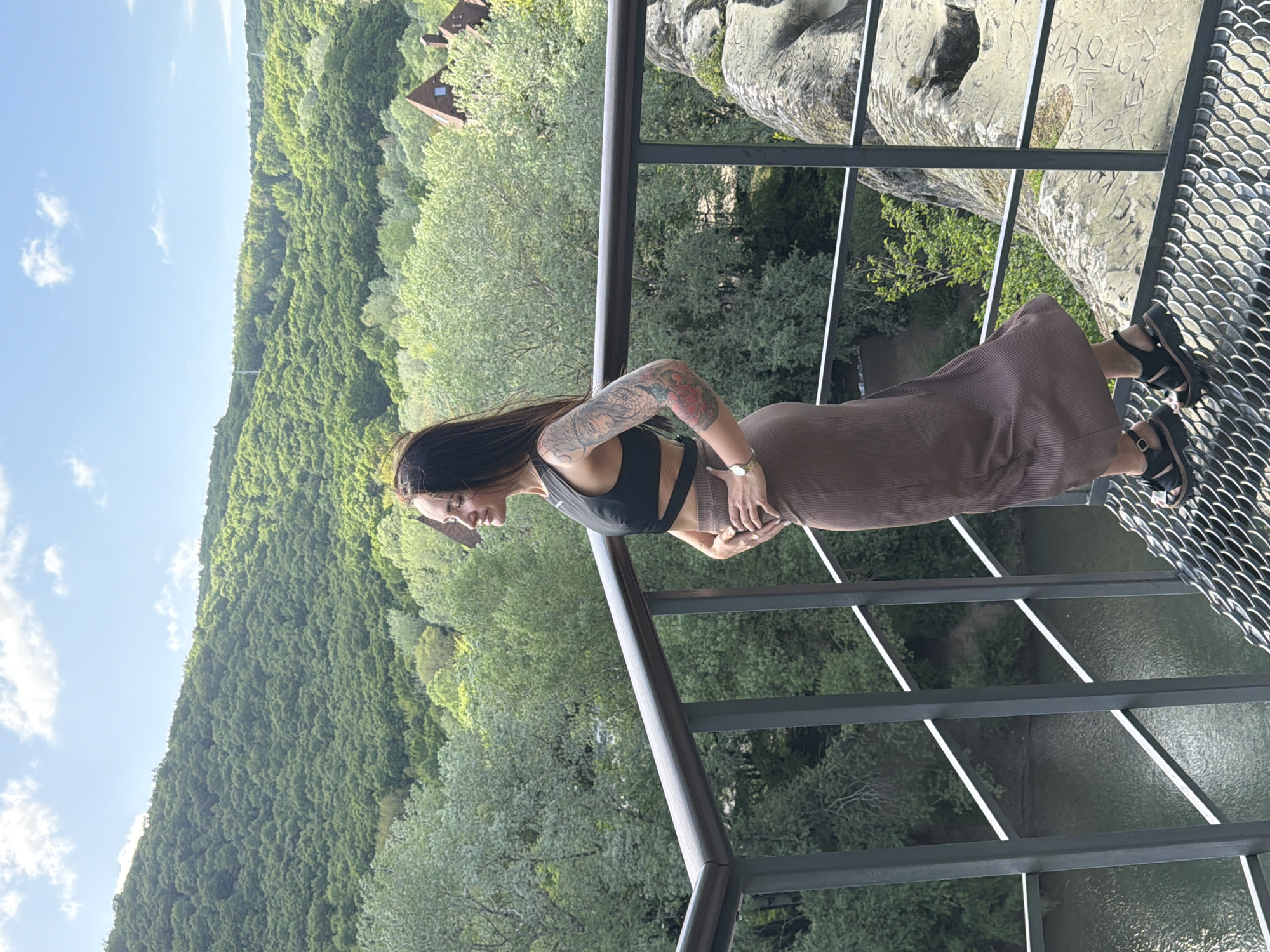 A full shot depicts a woman standing on a metal overlook platform, presumably in the city of Goryachiy Klyuch, Russia, with a heavily wooded valley and rock formations visible in the background. She has long dark hair and is wearing a black sports bra and a brown skirt. Her arms are positioned at her waist. Visible tattoos adorn her left arm. She is also wearing black sandals. The overlook has a metal railing. Through the metal grate of the overlook, a river and the forest floor are visible. The sky is partly cloudy with a light blue hue. In the background, there are several buildings among the trees.