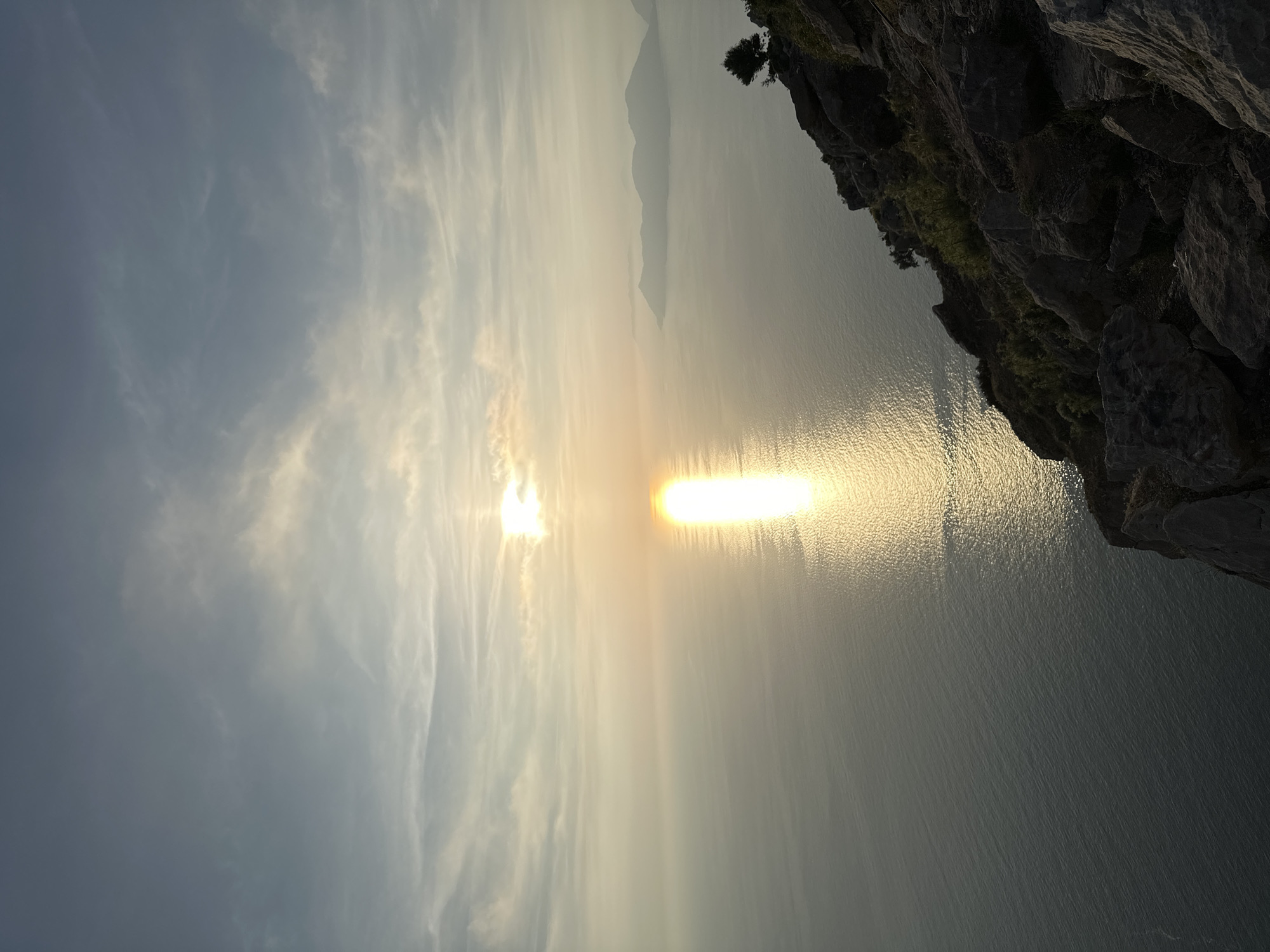 The image captures a serene sunset or sunrise over a vast body of water, likely the sea, viewed from a high vantage point along a rocky coastline in Fethiye, Turkey. The sun, a brilliant, glowing orb, is positioned low on the horizon, casting an intense, elongated golden reflection that shimmers across the water towards the viewer.
The sky is a blend of soft blue and wispy, streaky clouds, which are subtly illuminated by the warm, golden light of the sun, particularly towards the horizon. The atmospheric light suggests either early morning or late afternoon, with a gentle, hazy quality.
In the foreground, a dark, rugged cliff face occupies the right side of the frame. Its rough texture and shadowy crevices are visible, with hints of sparse green vegetation clinging to its surface, including a small tree or bush near the top edge. The water below the cliff also catches some of the golden light.
Across the expansive water, faint, hazy silhouettes of distant mountains or landmasses are visible, adding depth to the horizon. The water itself appears calm, with subtle ripples catching and reflecting the sun's brilliance. The overall scene evokes a sense of peace and natural grandeur. There is no visible text in the image.