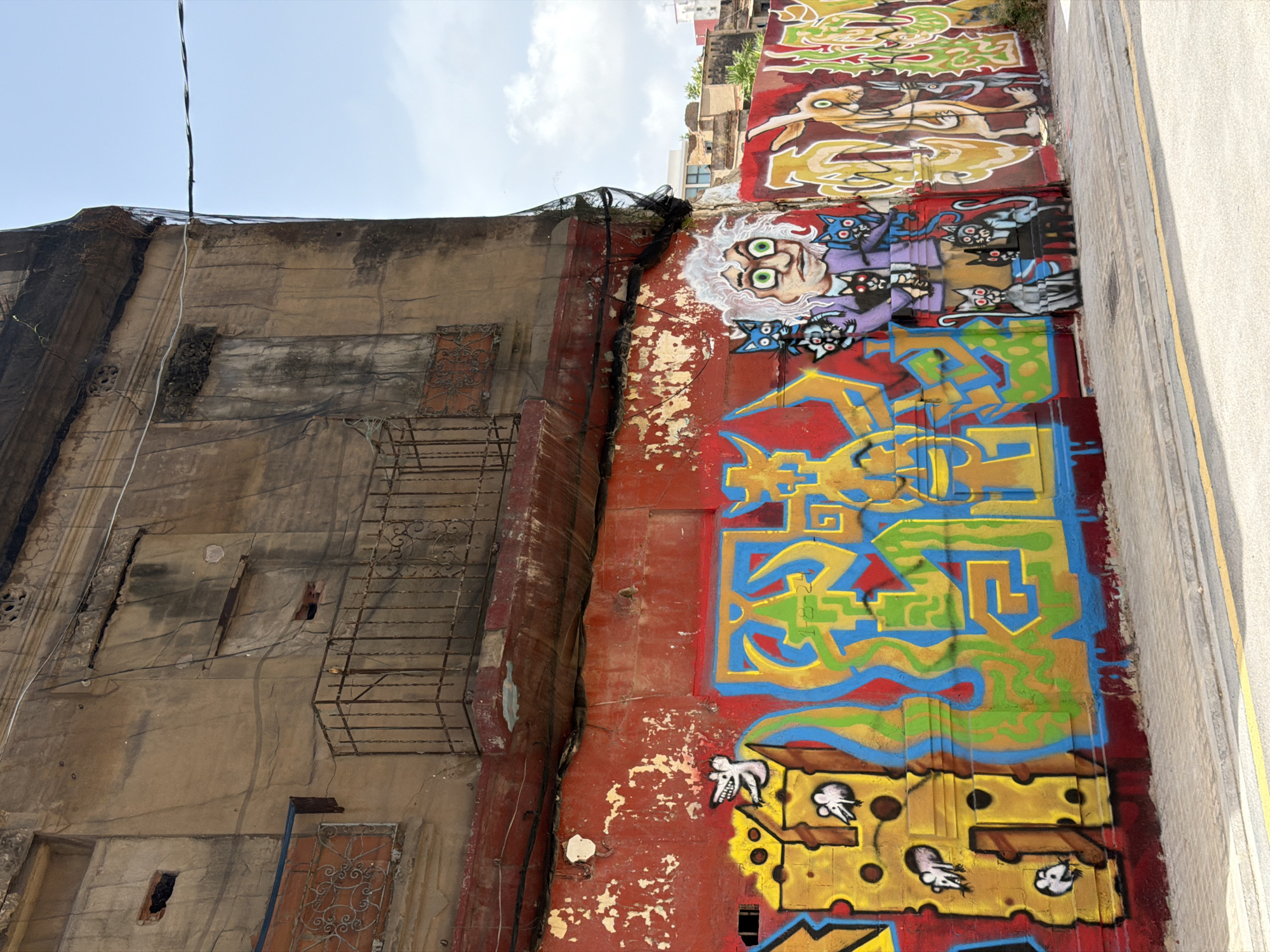 The image shows a close-up, low-angle view of a graffiti-covered wall and an adjacent building. The wall on the left is weathered and appears to be old, with patches of discolored concrete and exposed rebar in some areas. There are also remnants of what looks like a metal fire escape or support structure attached to this wall, and a piece of black netting draped over the top edge. The wall on the right is vibrantly covered in graffiti.
The graffiti on the right wall depicts several figures and designs. Near the top, there's a stylized depiction of a person with bright green eyes and wild white hair, surrounded by several blue cats with red eyes. Below this, more abstract, colorful graffiti dominates, featuring geometric shapes and wavy lines in blues, greens, yellows, and reds. At the bottom of the graffiti wall, there's an illustration of what appears to be Swiss cheese with cartoonish mice peeking out.
The scene is set outdoors, likely during the daytime, as indicated by the bright sky and shadows. The weather appears to be clear and sunny. The overall impression is one of urban decay juxtaposed with vibrant street art, suggesting a location in a city with a lively artistic scene. No discernible text is visible in the graffiti.