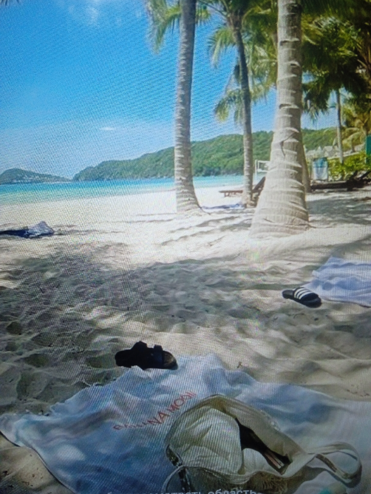 The image depicts a tropical beach scene with palm trees, sand, and the ocean. There are several objects on the sand, including a pair of flip-flops and a towel with some text that is partially visible. In the background, there are hills covered in lush greenery and a clear blue sky. The scene appears to be a peaceful and relaxing setting. There is no information available about this scene being located in Chernivtsi, Ukraine, as Chernivtsi is an inland city and not known for having tropical beaches.