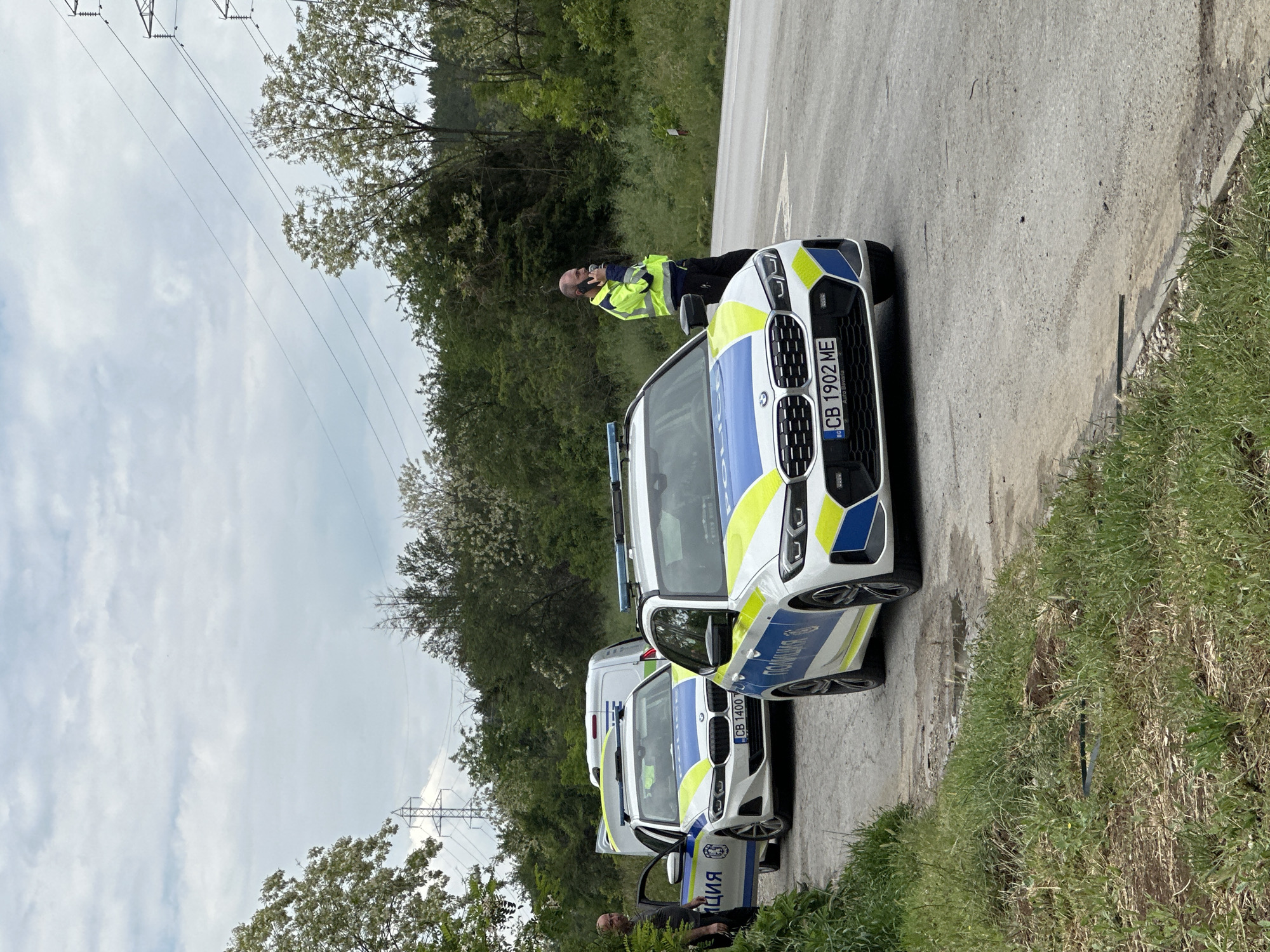 The image depicts a roadside scene in Ledenik, Bulgaria. Two Bulgarian police vehicles, a white BMW sedan with license plate CB 1902 ME and a white van with license plate CB 1400, are parked on the edge of a paved road. A male police officer in a yellow and black high-visibility jacket is standing next to the BMW sedan, holding a communication device near his ear. Another individual is visible near the van, and the open driver's side door reveals interior details. The surrounding environment includes green vegetation, trees, and power lines overhead, suggesting a rural or semi-rural location. The sky is overcast.