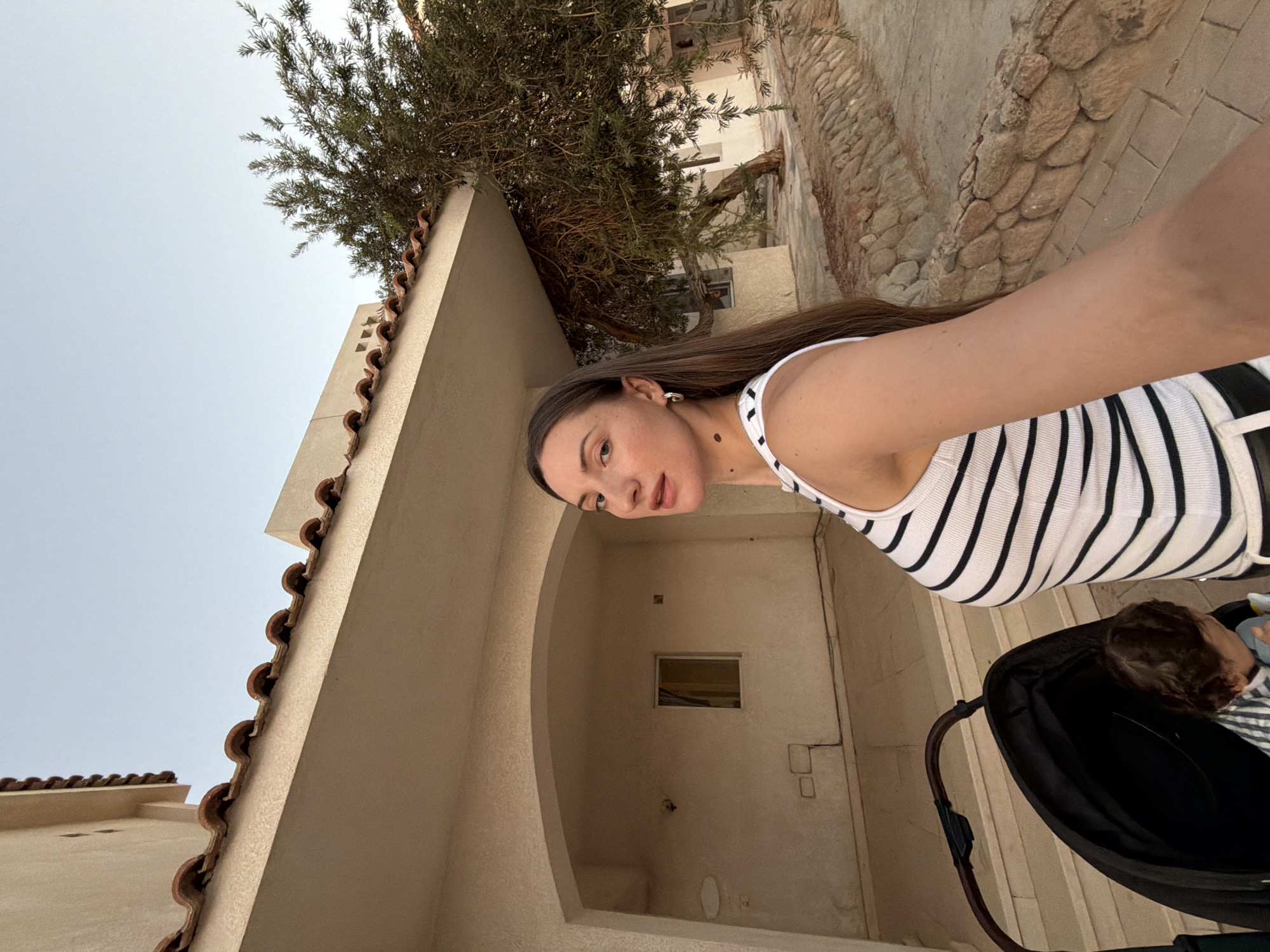 In Sharm El Sheikh, Egypt, a woman with long brown hair stands in front of a beige building with a red-tiled roof and an arched entrance. She wears a black and white striped tank top. To her left, a black stroller is visible, with a baby inside wearing a striped outfit. A tree with dense green foliage is situated to her right, with a stone border separating the paved walkway from a sandy area. The sky above is clear and light blue, indicating daytime.