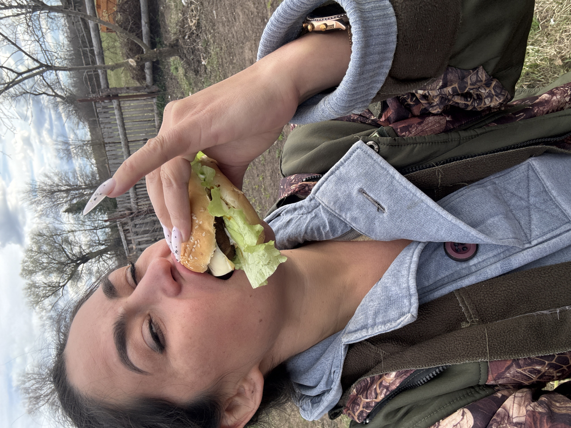 A woman is eating a hamburger outside. The woman has dark hair and is wearing a gray shirt with a brown and green camo jacket. Her nails are long and pointed. The hamburger has lettuce and a sesame seed bun. The background shows a wooden fence and trees, suggesting an outdoor setting potentially in a rural area near Кемуль, Russia.
