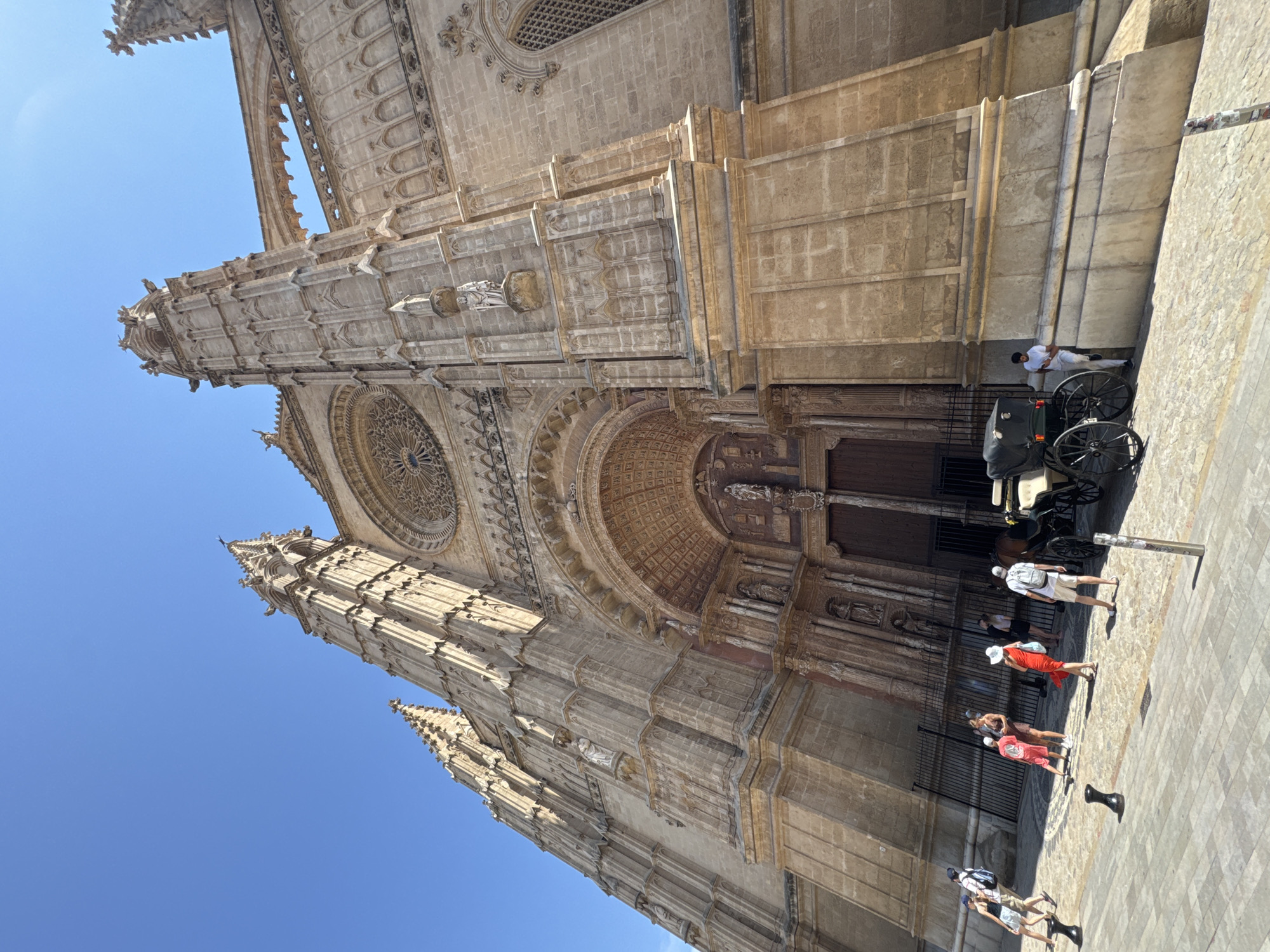 This is a low-angle, wide shot of the Palma Cathedral (La Seu) in Palma de Mallorca, Spain, under a clear blue sky. The grand Gothic cathedral, constructed of light-brown stone, dominates the frame, with its intricate facades, tall spires, and a large rose window visible. In the foreground, a cobblestone plaza is occupied by several tourists and a horse-drawn carriage.
Specifically, the scene depicts:
* **Location:** Palma Cathedral (La Seu), Palma de Mallorca, Spain.
* **Architecture:** The cathedral's imposing Gothic architecture is the primary subject. Details include carved stone facades, flying buttresses, pointed arches, spires reaching towards the sky, and a prominent rose window above the main entrance. Gargoyles are visible on the upper sections of the spires.
* **People:** Several individuals are present in the plaza. On the left, a group of tourists are walking. Further to the right, a person in a red dress and a white hat is standing, and another person in a white shirt and shorts with a backpack is facing away from the camera. A man in a white shirt and pants is leaning against a carriage.
* **Objects:** A dark-colored horse-drawn carriage with a black canopy is parked on the right side of the plaza. Black bollards are placed on the cobblestone.
* **Environment:** The scene is set in a sunlit outdoor plaza with a textured cobblestone surface. The sky is clear and bright blue. The angle of the shot emphasizes the height and grandeur of the cathedral.
The overall impression is one of a popular tourist destination on a bright, sunny day.
