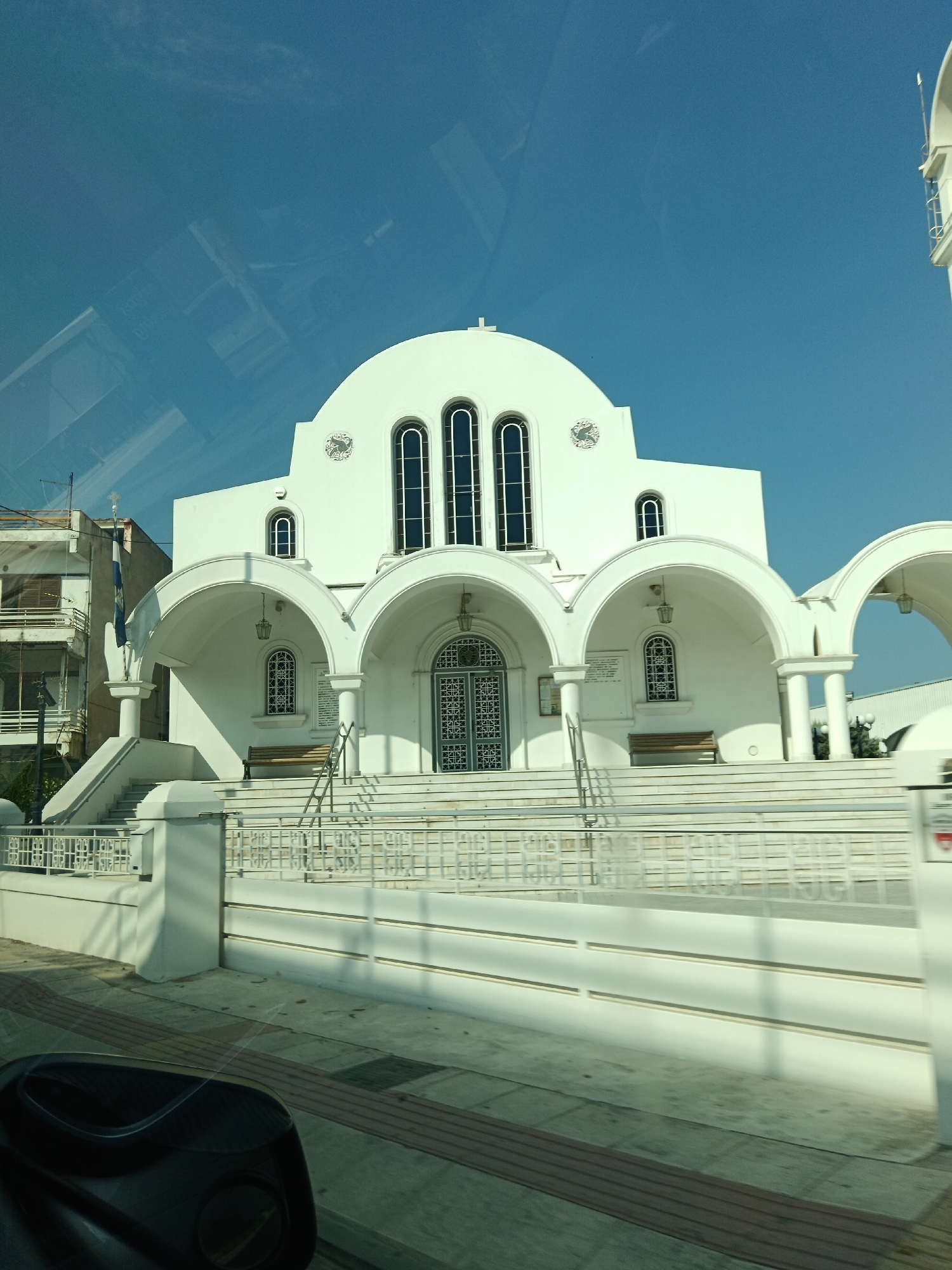 The image shows the exterior of a white, domed church with arched porticos and classical architectural elements. The church has tall, arched windows with dark frames and decorative metal grilles on some of the lower windows. A set of wide, white stairs leads up to the main entrance, with white railings on either side. There are benches on either side of the entrance. To the left of the church, a multi-story building with balconies is visible. A Greek flag is flying from a flagpole. The sky is clear and blue. The foreground shows a sidewalk and the edge of what appears to be a car's interior, suggesting the photo was taken from a vehicle. The specific location is the Municipal Unit of Velos, Greece, and the church is likely the Church of St. Nicholas in Velos.