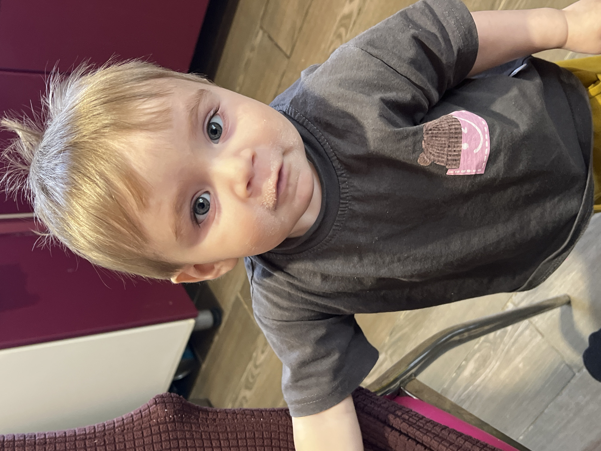 An indoor portrait captures a young child, likely a toddler, with light blonde hair and striking blue or gray eyes, looking directly at the camera. The child's face, particularly around the mouth and cheeks, is smudged with a light-colored, dried substance, suggesting they have recently finished eating.
The child is dressed in a dark grey, short-sleeved t-shirt. Adorning the right side of the shirt is a distinctive appliqué. This design consists of two parts: a brown, textured element resembling a patterned fabric, and an adjacent pink section that features a simple white line drawing of a smiling face. A glimpse of yellow clothing, possibly pants or shorts, is visible towards the bottom right.
The setting appears to be a domestic interior, possibly a kitchen or dining area in Noviy, Russia. The background on the left reveals a piece of dark red or burgundy furniture, such as a cabinet, situated above a white surface. The floor, visible towards the top right and bottom right, is made of light-colored wood or wood-laminate. In the bottom left, a dark red, textured fabric, possibly a blanket or part of a cushion, is present, and a shiny metal chair frame can be seen partially on the right side.
The child's expression is neutral and curious, with wide-open eyes. Their hair is somewhat tousled, with several strands standing upright. The image captures a candid, everyday moment. The lighting suggests an indoor environment, but the specific time of day or external weather conditions are not discernible. The only discernible text is the smiley face drawn on the shirt's appliqué.