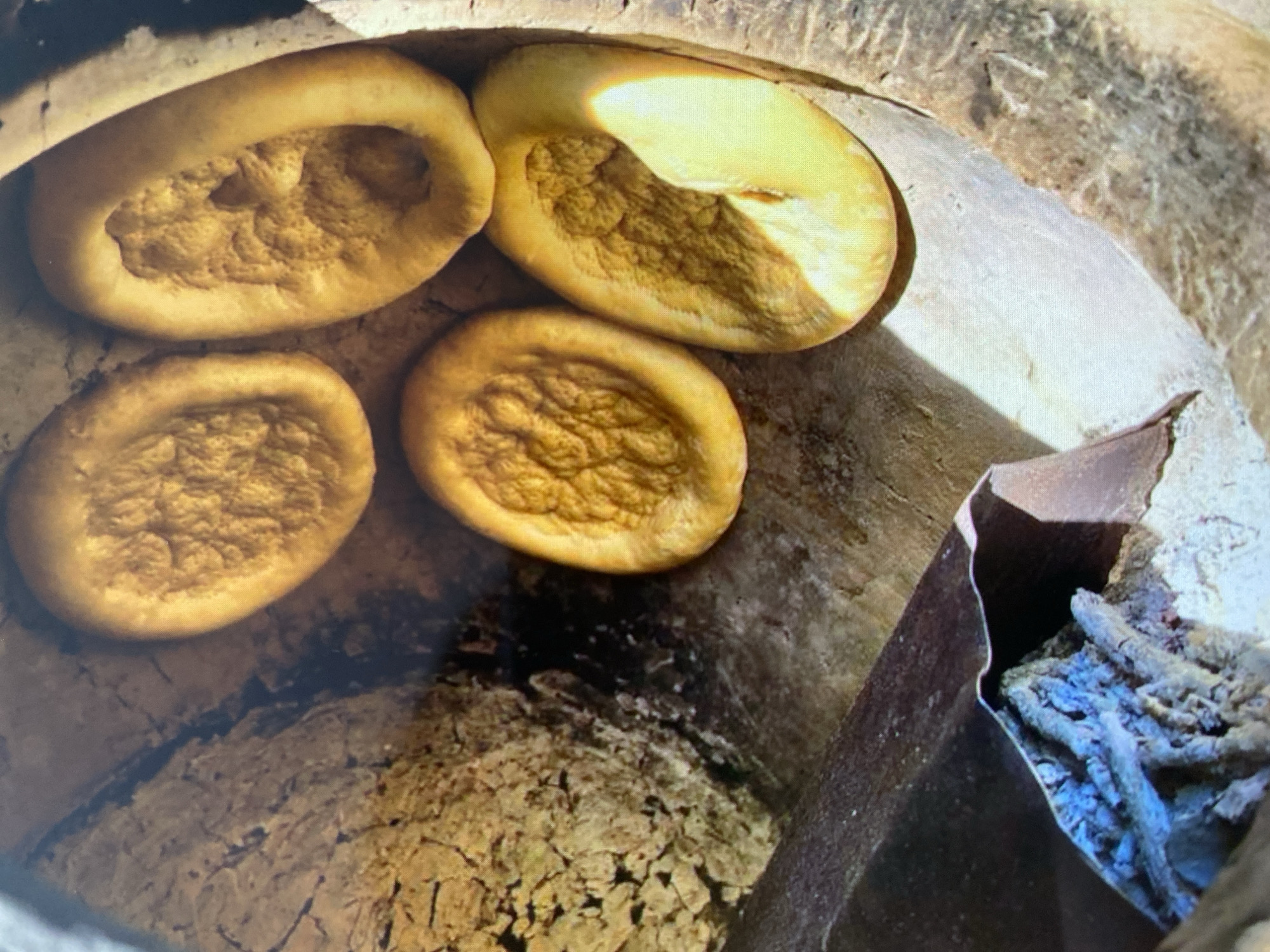 An overhead, close-up shot shows four pieces of golden-brown, baked flatbread in a tandoor oven. The tandoor is made of rough, light-brown clay with some darker, charred areas. To the left, a piece of dark metal with blue flames and ash is visible, suggesting a heat source. The flatbreads are irregular in shape, with a textured, bubbly surface. One piece, in the foreground, is larger and has a distinct indentation. The overall lighting is warm, highlighting the golden hues of the bread and the earthy tones of the oven.