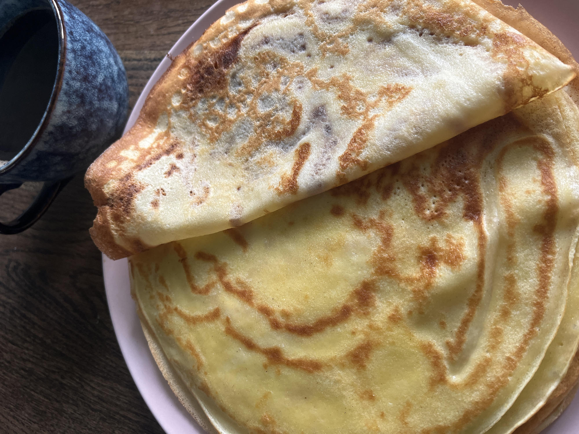 A close-up shot shows a stack of crepes on a pink plate next to a blue speckled mug, all sitting on a wooden surface. The top crepe is folded over, revealing a browned, slightly speckled surface. The stack appears to contain at least two or three crepes. The mug is partially filled with a dark liquid, possibly coffee or tea. The wooden surface has a dark, grainy texture. There is no information regarding the city of Krasnodar, Russia in the provided content.