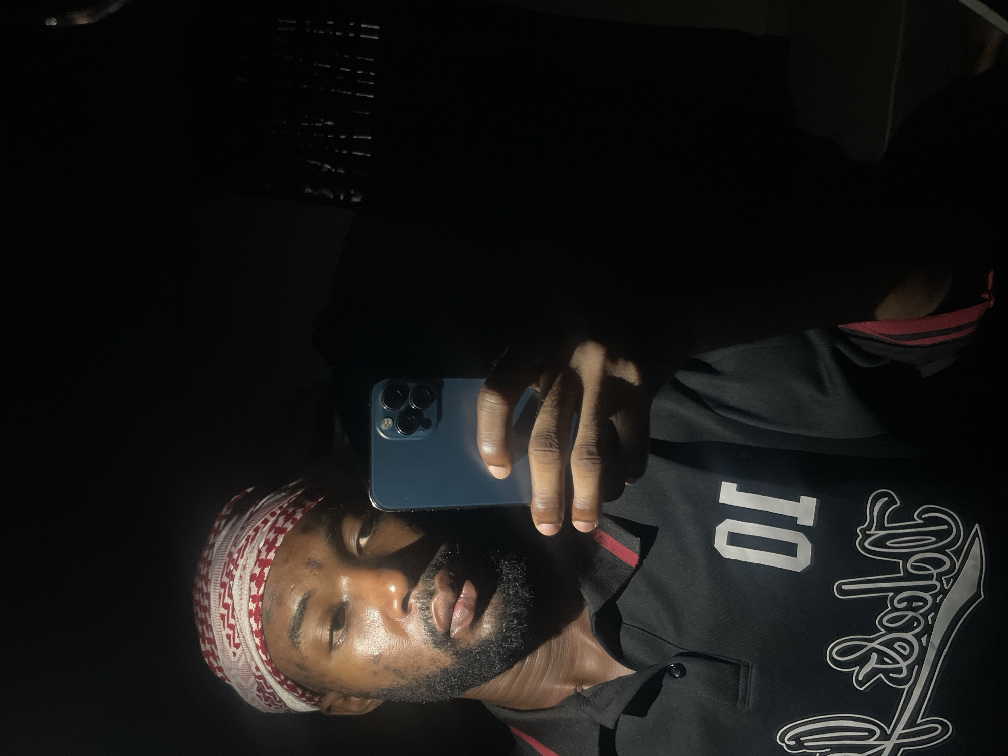 A man is taking a selfie with his phone. He is wearing a red and white patterned head covering and a black shirt with a white design and the number "10." The background is dark, but the man is illuminated by a light source. There is no location evidence in the image. Therefore the location in the city of Lokoja, Nigeria cannot be included.