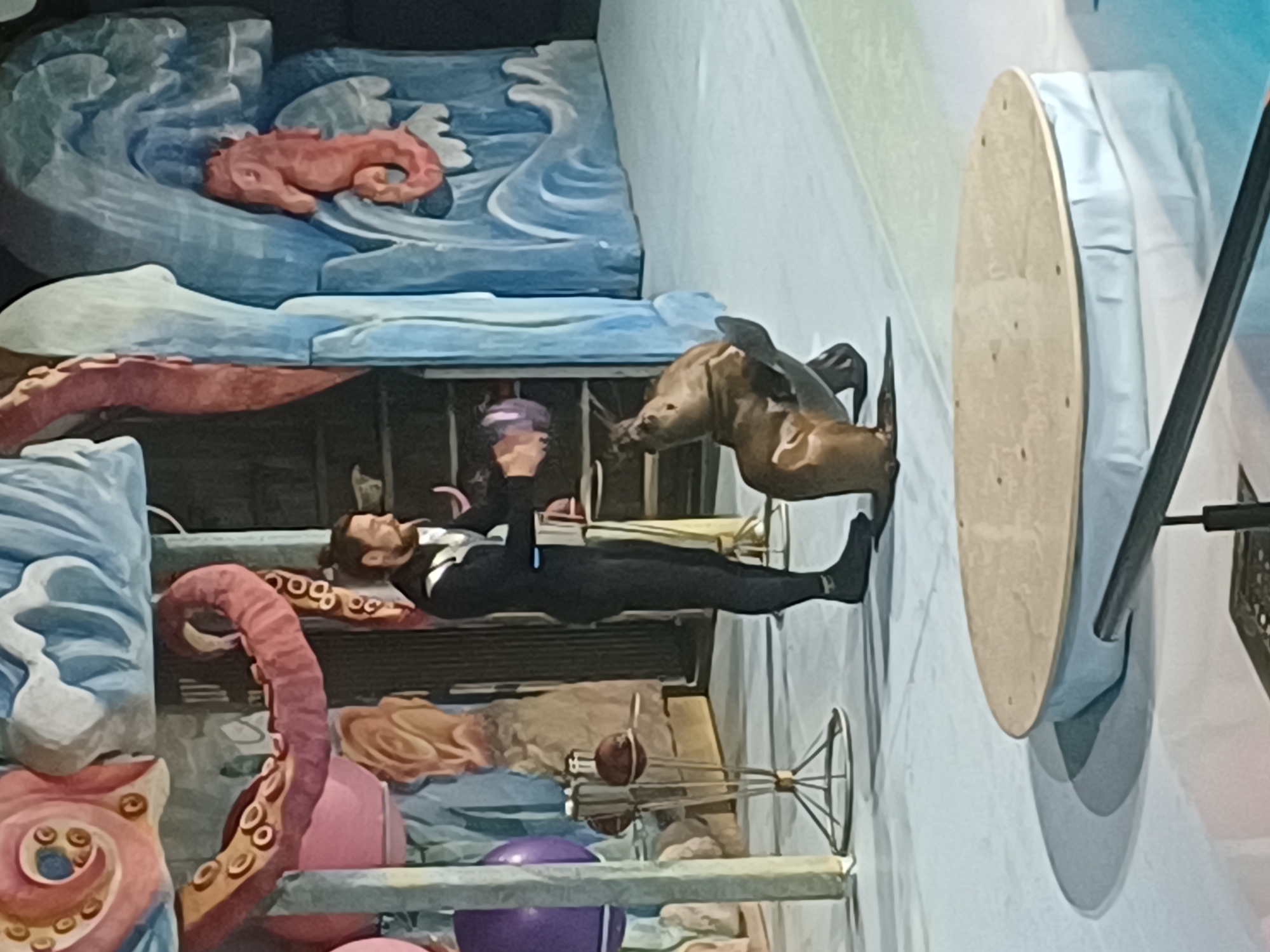 A male trainer in a black wetsuit interacts with a sea lion on a light-colored stage in what appears to be an aquatic-themed performance venue. The trainer, with a beard and dark hair tied back, stands with his left foot forward, holding an object in his gloved hands towards the sea lion. The sea lion is seated on its haunches, looking up at the trainer, with its front flippers raised. The background features a large, colorful mural depicting underwater elements, including pink octopus tentacles, blue waves, and a coral-colored seahorse. In the foreground, a circular wooden platform with a light blue skirt is visible. The floor of the stage is a smooth, light surface. The overall impression is of a live animal training demonstration or performance. The location is not specified to be Novosibirsk, Russia.