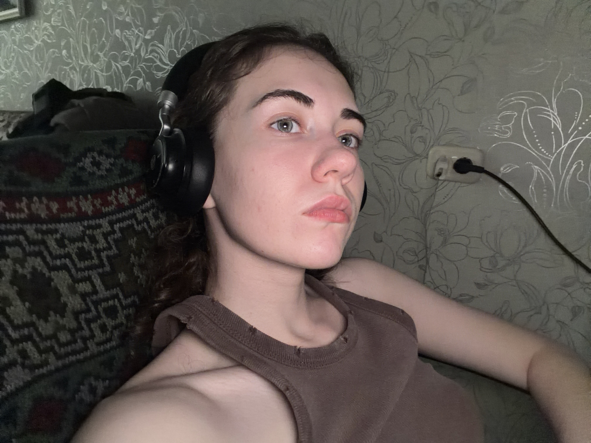 The media file shows a young woman with light skin and brown curly hair, wearing black headphones. She is looking at the camera with a neutral expression, her eyes are light blue. She is wearing a brown tank top. The background appears to be a room with floral patterned wallpaper and a patterned sofa. There is an electrical outlet on the wall with a black plug inserted. The lighting suggests it is nighttime or indoors in low light. The overall mood is casual and relaxed.