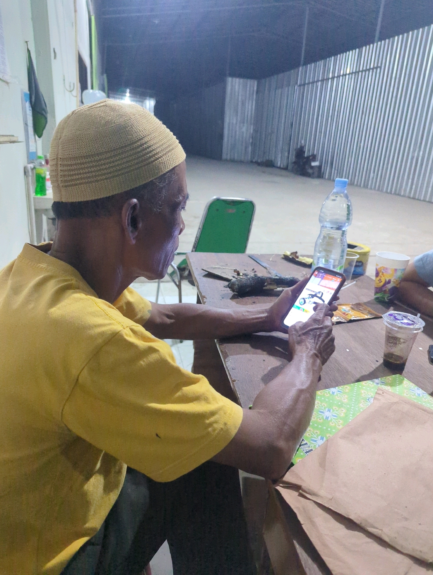 An Indonesian man wearing a yellow t-shirt and a brown skullcap is seated at a wooden table, looking at his smartphone. The man's right hand is holding the phone, and his left hand rests on the table. The table is cluttered with various items, including a bottle of water, a disposable cup with a dark liquid, and some brown paper bags. In the background, a corrugated metal fence and a green plastic chair are visible. The scene appears to be outdoors, possibly in a backyard or a common area, given the open space and the materials used for the fence. The lighting suggests it might be evening or nighttime. The location is identified as Batu Belaman, Indonesia.