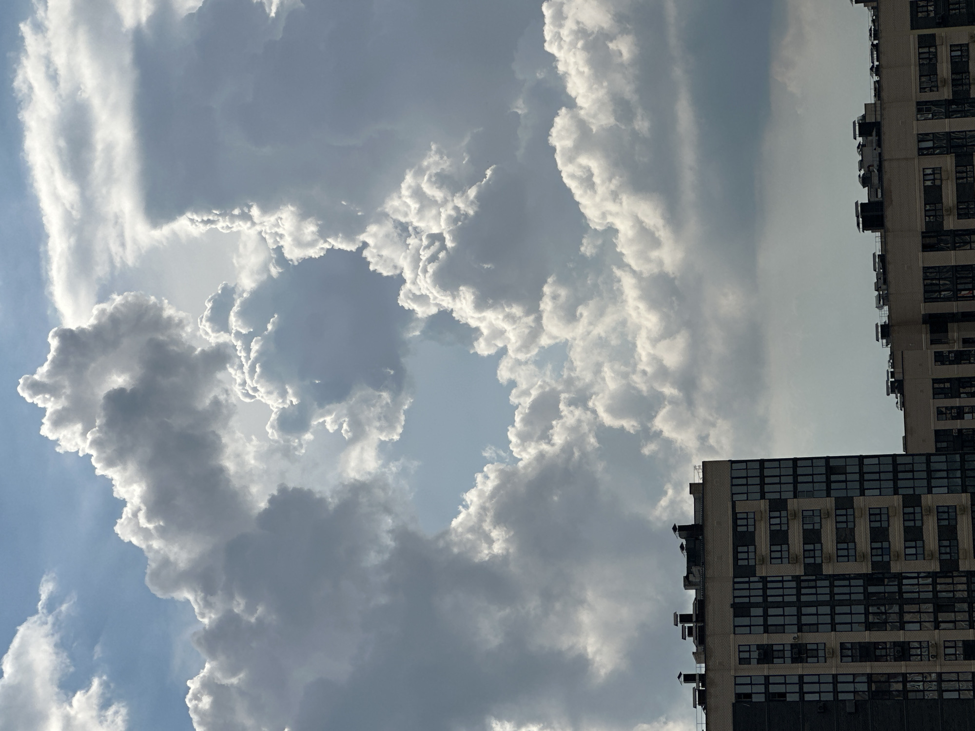 The image captures a dramatic sky filled with large, textured clouds over a city. The clouds vary in color from bright white where illuminated by sunlight to dark gray in shadowed areas, suggesting a dynamic weather system. A gap in the clouds reveals a bright blue sky.
In the foreground, the tops of two modern apartment buildings are visible. The building on the left is taller and features a facade of dark windows interspersed with lighter colored panels, creating a grid-like pattern. Several ventilation shafts or chimneys are visible on the roof of this building. To the right, the roofline of a second, lower building is visible, also with ventilation units.
The scene is shot from a high vantage point, looking upwards towards the sky. The overall impression is one of a partly cloudy day with impressive cloud formations over an urban landscape. This image is identified as being taken in Krasnodar, Russia.
