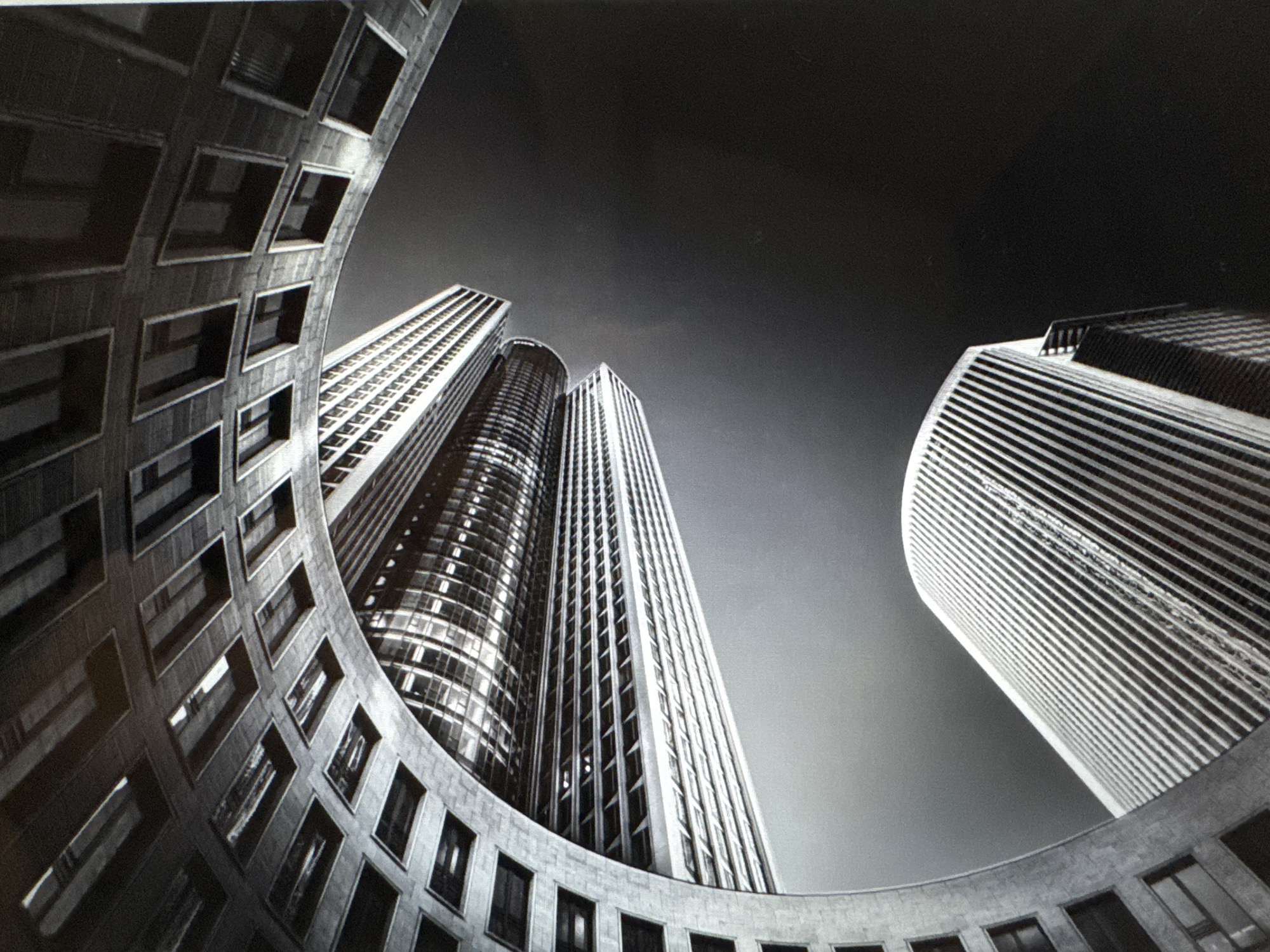 The image is a striking monochromatic architectural photograph, captured from a low-angle perspective looking upwards at a cluster of modern buildings against a stark sky.
In the foreground, a prominent curved building with a textured, possibly stone or concrete facade, frames the lower and left portions of the composition. This building features numerous square windows arranged in a grid pattern, some reflecting light. Its sweeping curve guides the eye towards the central elements.
Beyond this foreground structure, several towering skyscrapers dominate the mid-ground. One central, dark, and highly reflective building displays a smooth, curved glass facade, catching highlights. To its right, another tall building exhibits strong, uniform vertical lines along its entire height. On the far right, partially visible, a third skyscraper rises, characterized by distinct horizontal banding across its surface.
The dramatic perspective emphasizes the height and grandeur of the urban landscape. The sky transitions from a lighter gray near the buildings to a deep, dark gray or black, particularly towards the upper right, creating a high-contrast and slightly dramatic atmosphere. The scene is devoid of people or vehicles. There is no visible text. The overall impression is one of sleek, modern urban architecture, highlighted by the black and white aesthetic which accentuates shapes, lines, and textures.