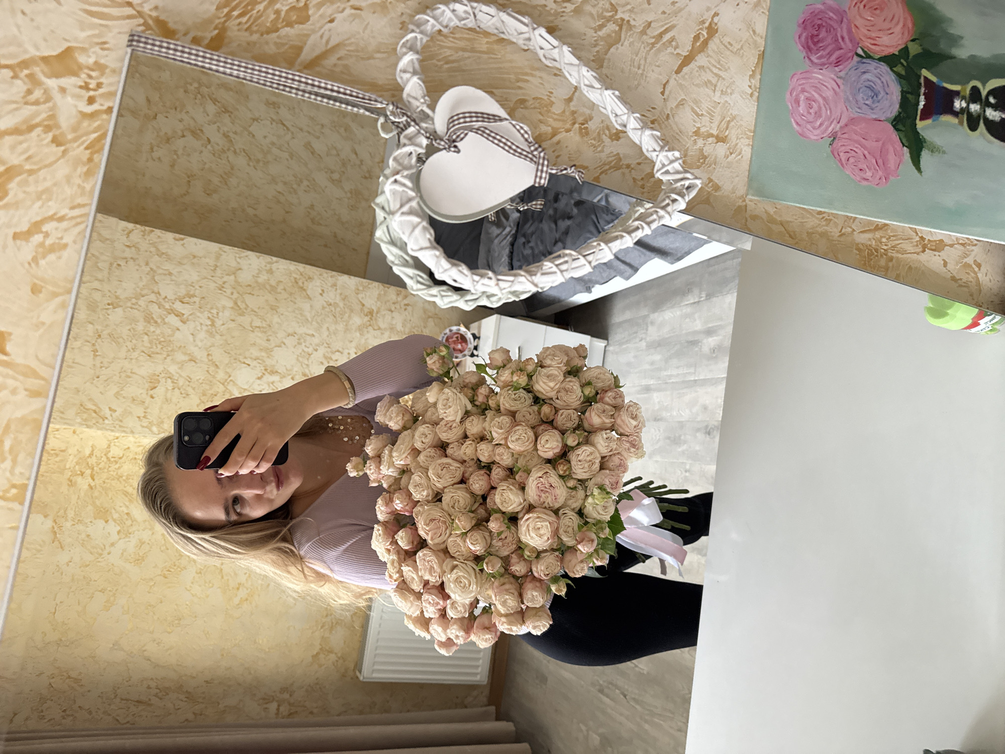 A reflection in a mirror shows a woman holding a bouquet of pink roses. The mirror hangs on a wall with textured beige wallpaper. A decorative heart-shaped wreath is attached to the wall above the mirror. A painting of colorful roses hangs on the wall to the right of the mirror. The woman is taking a photo of herself with her phone. She is wearing a light purple top and black pants. The room appears to be indoors. The location and city are unknown.