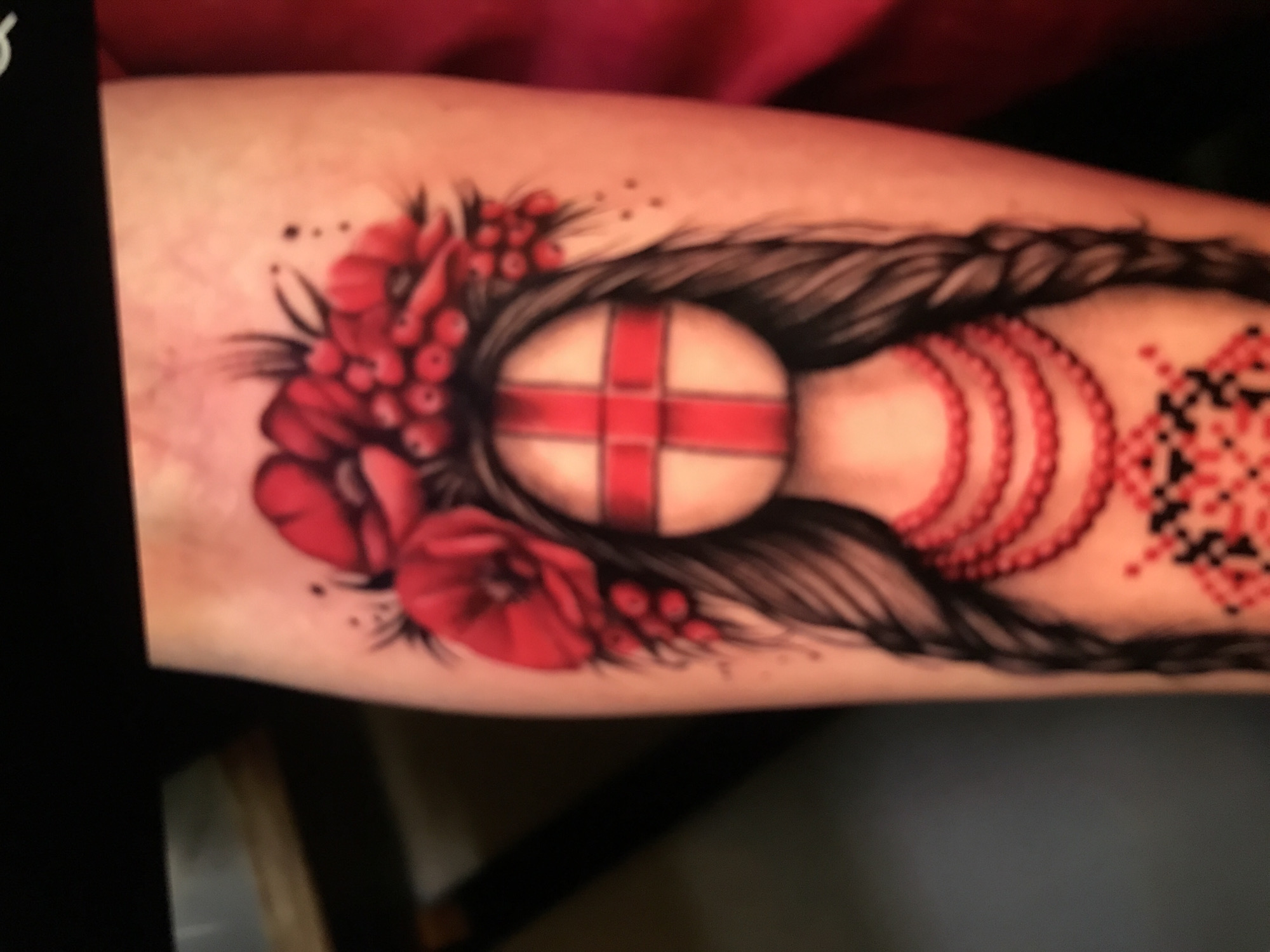 The image shows a tattoo on a forearm. The tattoo depicts a central oval shape with a red cross on a white background. Surrounding the oval are floral elements in red and black, including poppies and berries. Black braids of hair frame the central oval. To the right, there are red beaded strands and a black and red geometric pattern. The scene does not depict the city of Donji Pobori, Montenegro.