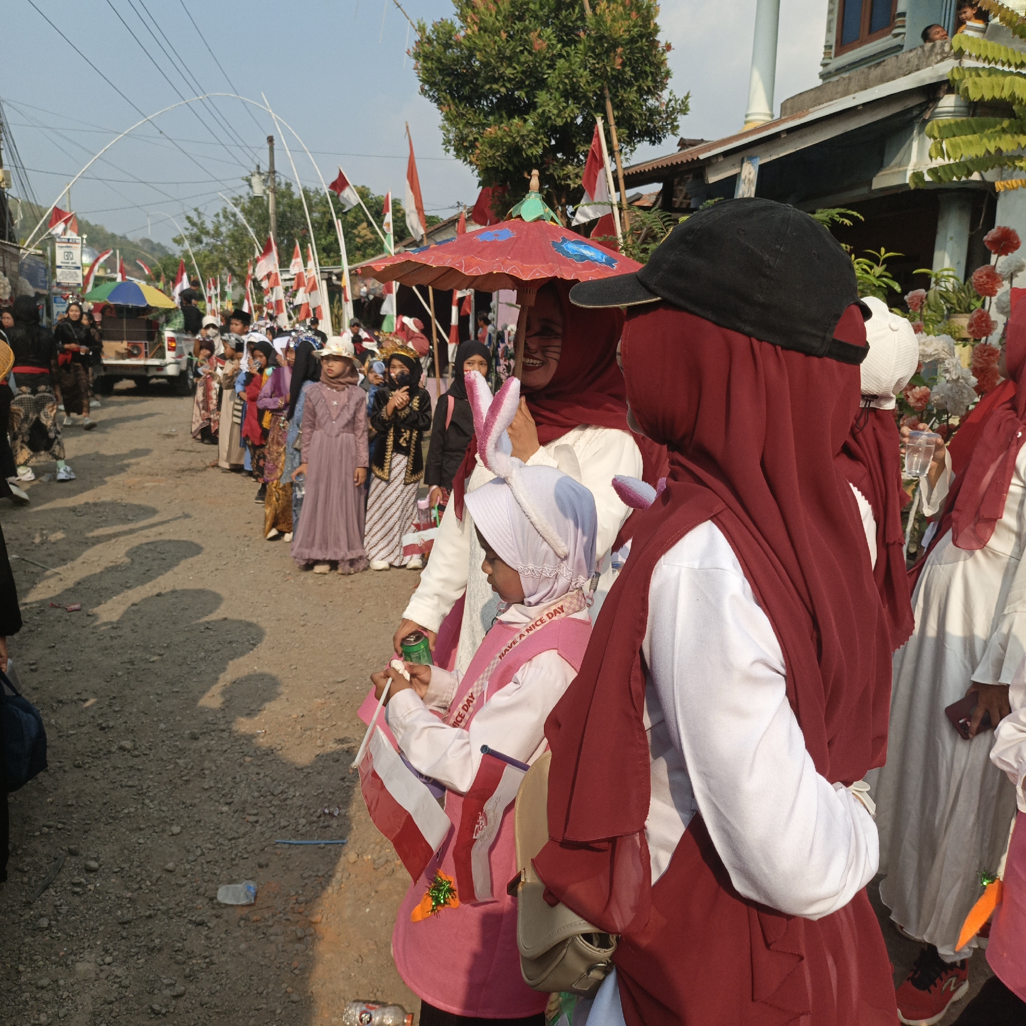 The image captures a festive gathering on a sunlit, gravelly street in Tanahbaya, Indonesia. A procession is underway, indicated by a line of people, many adorned in traditional attire and headscarves. Indonesian flags, red and white, are prominently displayed on poles and held by individuals, suggesting a national celebration.
In the foreground, two individuals are the primary focus. The one on the left, facing away from the camera, wears a dark baseball cap and a maroon headscarf. Next to them, a young child in a pink tunic and white headscarf, complete with rabbit ears, holds small Indonesian flags and a green can. The child also wears a pink sash with the words "HAVE A NICE DAY". Behind them, another person in a maroon headscarf and white clothing is partially visible.
Further into the scene, a diverse group of people forms a procession. Some wear formal attire, while others are dressed more casually. Children are interspersed throughout the crowd, many holding flags. A red and white umbrella, decorated with colorful patterns, is positioned above some of the participants. A white pick-up truck, possibly part of the procession, is visible in the background.
The environment consists of a dirt and gravel road with scattered debris, including plastic bottles. Buildings with traditional Indonesian architecture, featuring wooden elements and exposed roof beams, line one side of the street. Overhead, a network of electrical wires crisscrosses the sky. The overall atmosphere suggests a community event, likely a parade or celebration, with a strong sense of national pride.