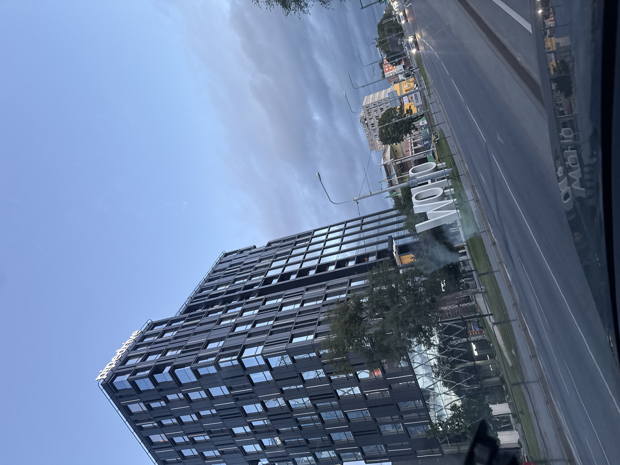 The image depicts a street scene in Tallinn, Estonia, at dusk. A modern, dark-colored office building with a reflective glass facade dominates the left side of the frame, bearing the illuminated "pipedrive" logo at its apex. In front of the building, a large, white, three-dimensional sculpture spelling "WOHO" is visible. The street in the foreground shows lanes of asphalt with lane markings. Cars are driving on the street, with headlights illuminated. In the background, other buildings, streetlights, and trees are present. The sky is overcast with shades of blue and gray, indicating evening. The scene appears to be captured from a moving vehicle, as suggested by the slight blur and angled perspective.