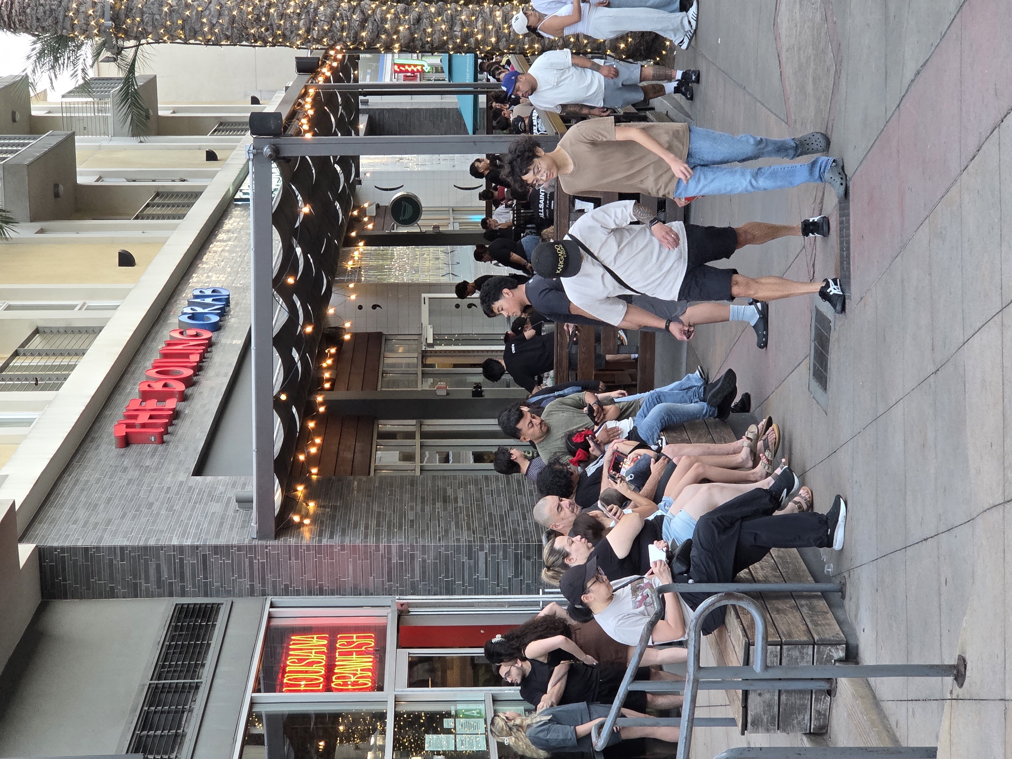 The image depicts an outdoor scene in front of a restaurant called "The Boiling Crab" located in Burbank, United States of America. A group of people are gathered outside the establishment. Several individuals are seated on a wooden bench, some engaged with their mobile phones, while others are conversing. Two young men are walking on the sidewalk in the foreground, one wearing a light-colored t-shirt and shorts, the other in a tan t-shirt and jeans. The restaurant's facade features a neon sign that reads "LOUISIANA CRAWFISH" in red lettering. The building is modern with multiple stories and balconies visible. A palm tree adorned with string lights stands to the right of the scene. The overall atmosphere suggests a casual dining environment on a pedestrian walkway.