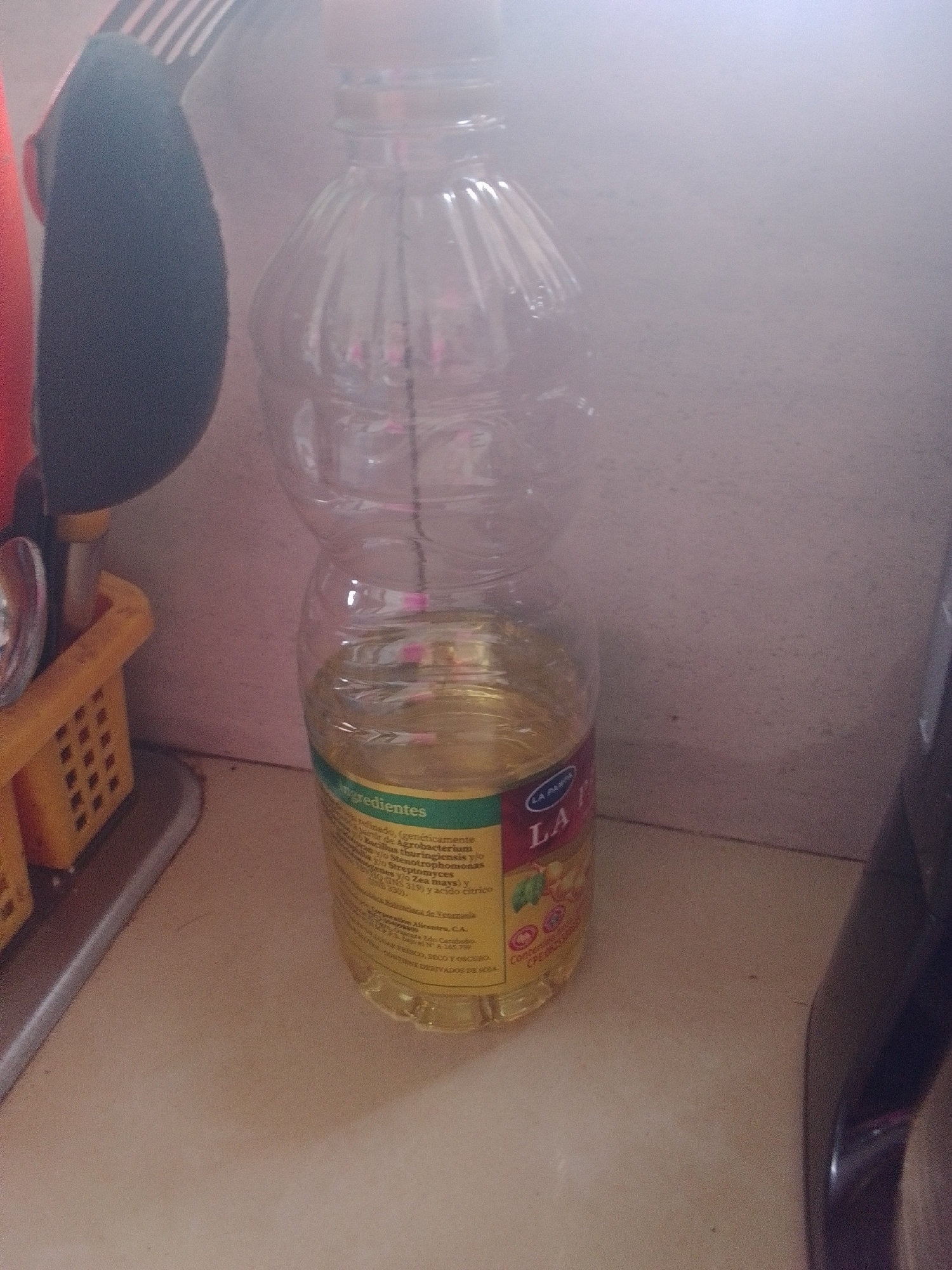 A bottle of cooking oil, likely La Parmesana brand, is positioned in the foreground of the image. The oil is a clear yellow liquid. A label on the bottle contains text in Spanish, indicating ingredients and origin. The bottle appears to be made of clear plastic and is partially filled. To the left of the bottle, a yellow plastic basket holds kitchen utensils, including what appears to be a whisk with black and white stripes. The background is a light-colored wall, possibly in a kitchen setting. The lighting is soft and diffused. There is no visible indication of the specific location within the city of La Tagua, Venezuela.