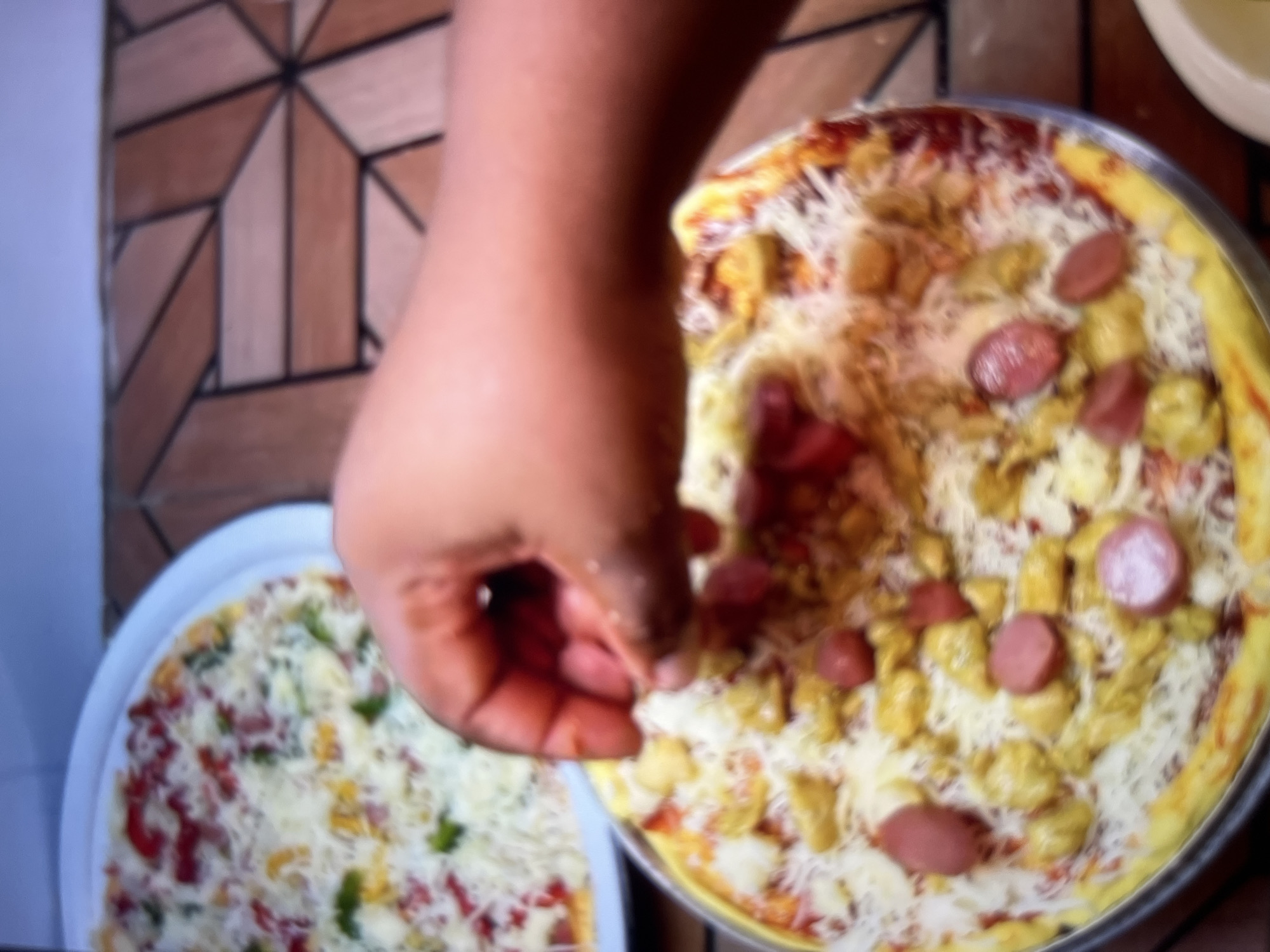An out-of-focus, overhead shot shows two pizzas. In the foreground, a pizza is being prepared. A dark-skinned hand is seen sprinkling toppings onto a pizza that is covered in cheese and some red sauce. The pizza has circular pieces of sausage and yellow toppings, possibly chicken or pineapple chunks, scattered across it. The pizza is in a metal pan. In the background, a second, slightly blurry pizza sits on a white plate. This pizza appears to have similar toppings, including red pieces, green pieces, and cheese. The pizzas are on a wooden table with a geometric pattern. The scene is indoors. This image is not specific to Abuja, Nigeria.