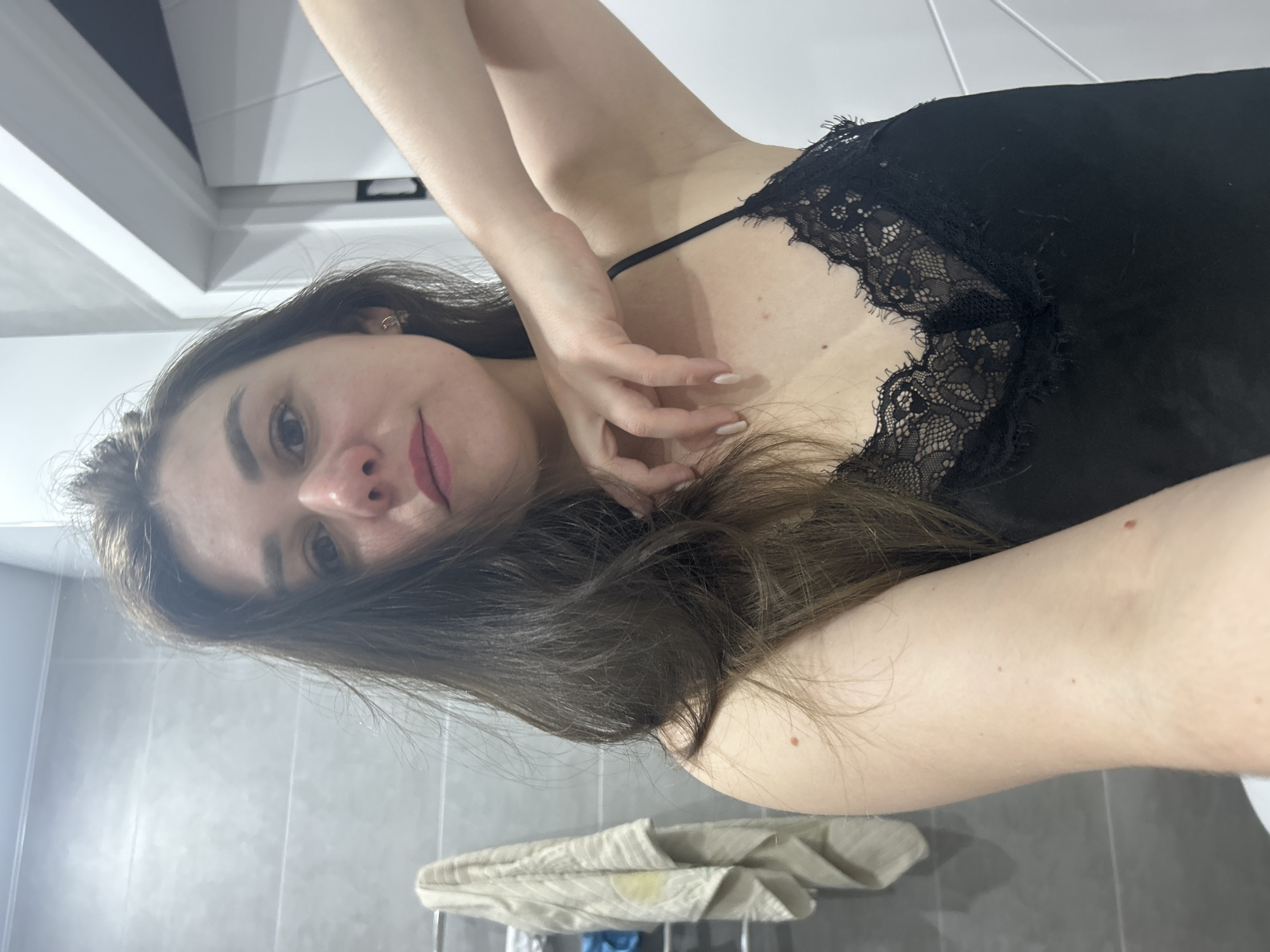 A woman is indoors, possibly in a bathroom, wearing a black camisole with lace trim. The background includes gray tiled walls and a towel rack with folded towels. The location is unknown, and there is no verifiable connection to Chaikovsky, Russia based on the visual information provided.