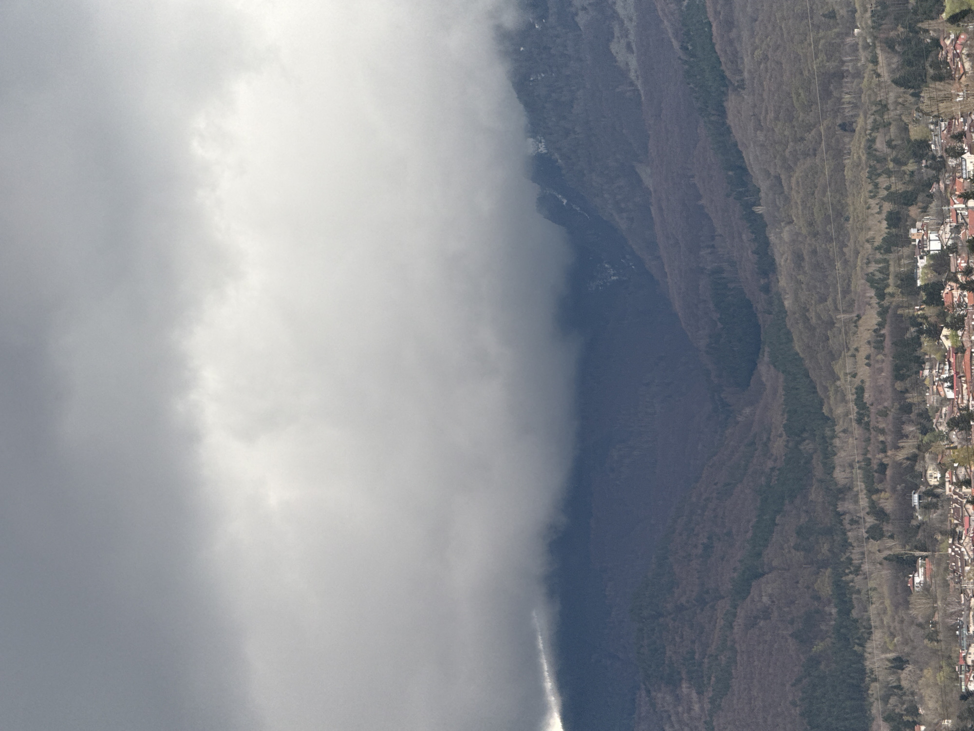 The image shows a view of a mountain partially obscured by heavy clouds. The mountain slopes are covered in trees, and at the base of the mountain is a densely populated area with buildings, likely a part of the city of Sofia, Bulgaria. The overcast sky dominates the left side of the image, giving the scene a gloomy atmosphere.