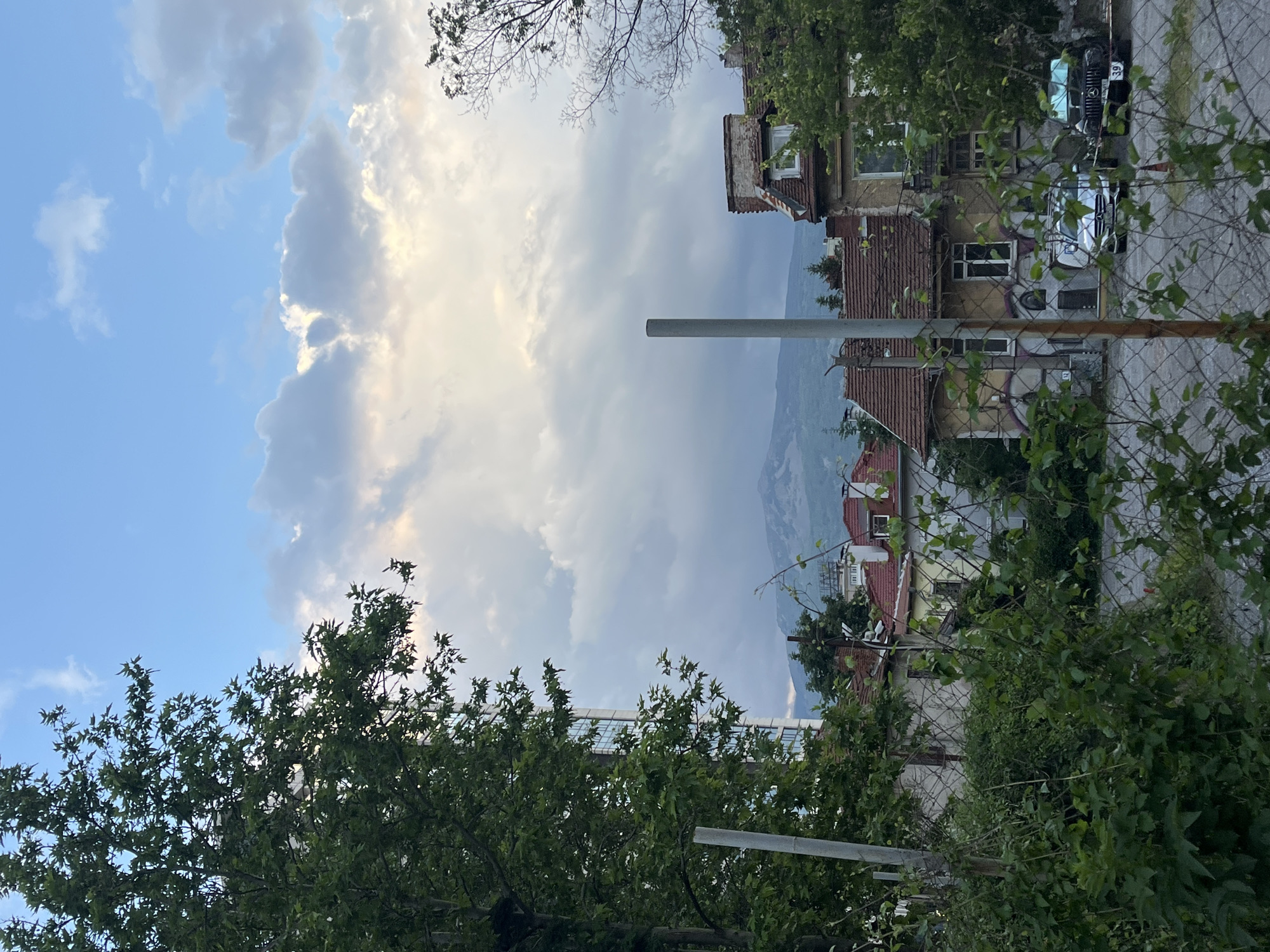 The image, oriented horizontally, depicts a layered view of an urban landscape in Sofia, Bulgaria, set against a backdrop of mountains and a dramatic sky. The upper portion of the frame is filled with a partly cloudy sky; clear blue is visible on the left, transitioning into large, voluminous grey and white clouds that show hints of sunlight, suggesting late afternoon or early evening.
In the foreground and mid-ground, dense green foliage from trees and bushes partially obstructs the view. A chain-link fence runs horizontally across the mid-section of the image, separating the viewer from the buildings and street beyond. Behind the fence, a paved street hosts at least two parked cars. A dark Mercedes-Benz sedan is clearly visible, with the number "39" on its license plate. Another lighter-colored car is partly seen beside it.
Several old, multi-story buildings with reddish-brown tiled roofs are present on the right side and central mid-ground. Their facades appear weathered, and one building features a large, colorful mural or graffiti design. A prominent grey pole or pillar stands vertically in the mid-ground. In the far distance, hazy blue mountains rise, forming a natural barrier behind the urban structures. No people are visible, and no specific activity is occurring.