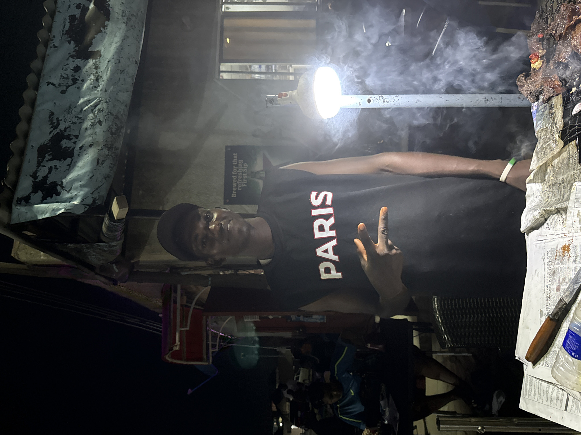 The image captures a nighttime scene at a street food stall in Abuja, Nigeria. A young man, wearing a black sleeveless shirt with "PARIS" printed in white, stands behind a grill. He is holding up two fingers in a peace sign. Smoke billows around him from the grill, which displays pieces of meat. To the left of the man, other people are visible, some seated at tables. The stall appears to be under a makeshift roof, illuminated by a bright lamp. Newspapers and a bottle of water are on the counter in the foreground. The overall environment suggests a bustling street food setting.