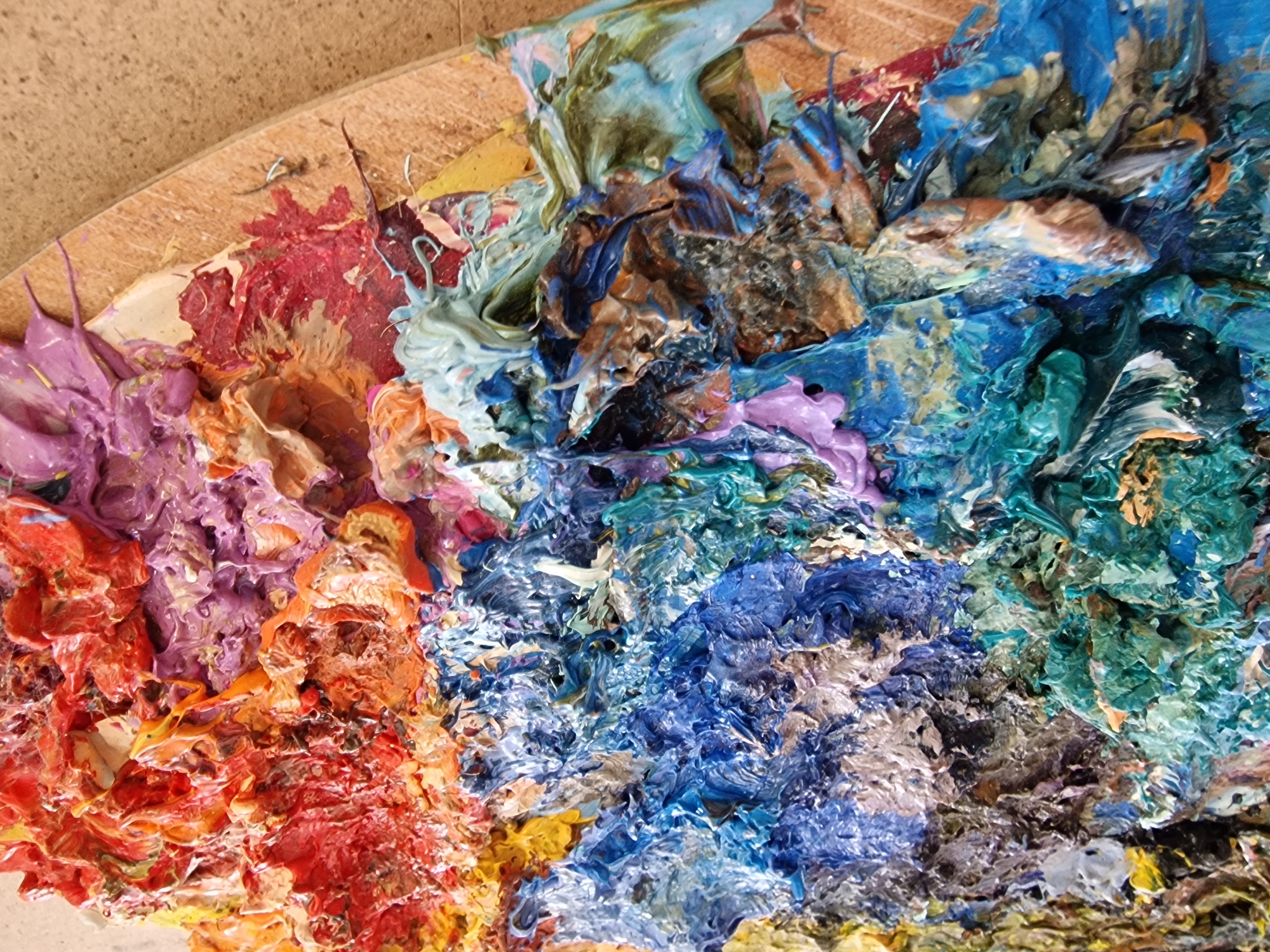 This image, captured in Veliko Tarnovo, Bulgaria, depicts a close-up, high-angle view of an artist's wooden palette covered in thick, impasto paint. The scene presents no individuals, actions, or direct interactions. The primary subjects are the heavily textured layers of paint and the underlying wooden palette, visible along the upper right edge.
The paint exhibits extreme dimensionality, with distinct peaks and valleys formed by a thick application. A broad spectrum of colors is present: vibrant reds, oranges, and yellows dominate the upper left quadrant. Adjacent to these are patches of purple and pink hues, mixed with lighter tones. The lower and central sections are primarily composed of various shades of blue and teal, interspersed with greens, whites, browns, and some grey and metallic-like streaks. The colors appear both distinct and partially intermingled, suggesting a history of mixing and application. The wooden palette edge is light brown with a visible grain, showing a curved perimeter. No other environmental details are discernible.