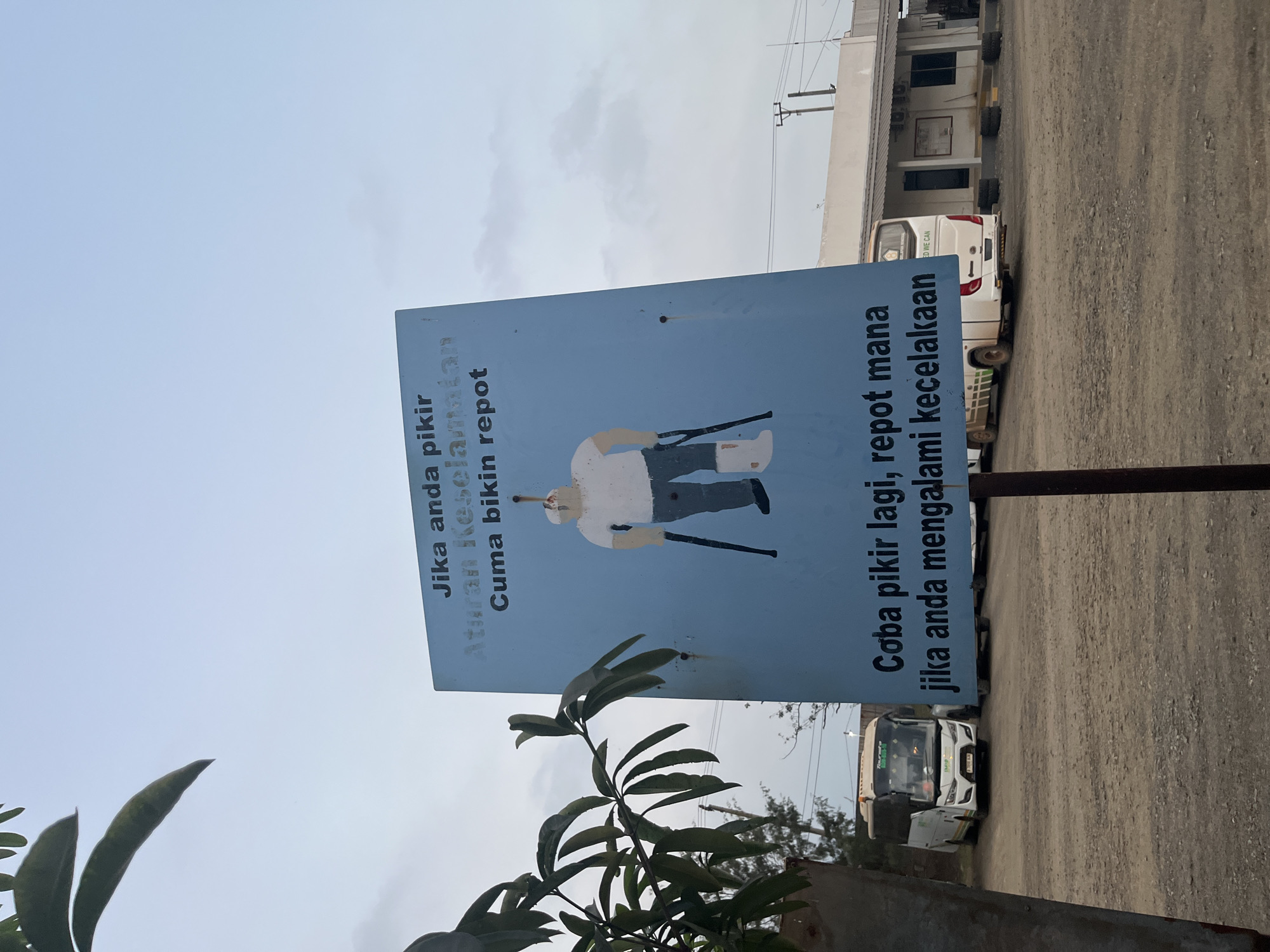 An outdoor, eye-level, medium shot captures a blue sign with a drawing and text, placed on a pole in what appears to be a parking lot or depot. The sign features a cartoon depiction of a person with a cast on one leg, using crutches, and an injury on their head, implied by a bandage or stain on their scalp. The text on the sign, in Indonesian, reads "Jika anda pikir Aturan Keselamatan Cuma bikin repot" above the drawing, and "Coba pikir lagi, repot mana jika anda mengalami kecelakaan" below it.
In the background, a white bus is parked to the left, and another white bus is visible further back to the right, partially obscured by a building. The building has a light-colored facade with dark-trimmed windows and a covered walkway. The ground is covered in gravel. Vegetation, including a branch with large leaves, is visible on the left side of the frame, framing the sign and obscuring part of the sky. The sky itself is overcast and pale. The overall impression is a reminder about safety, emphasizing the greater inconvenience of accidents compared to following safety rules.