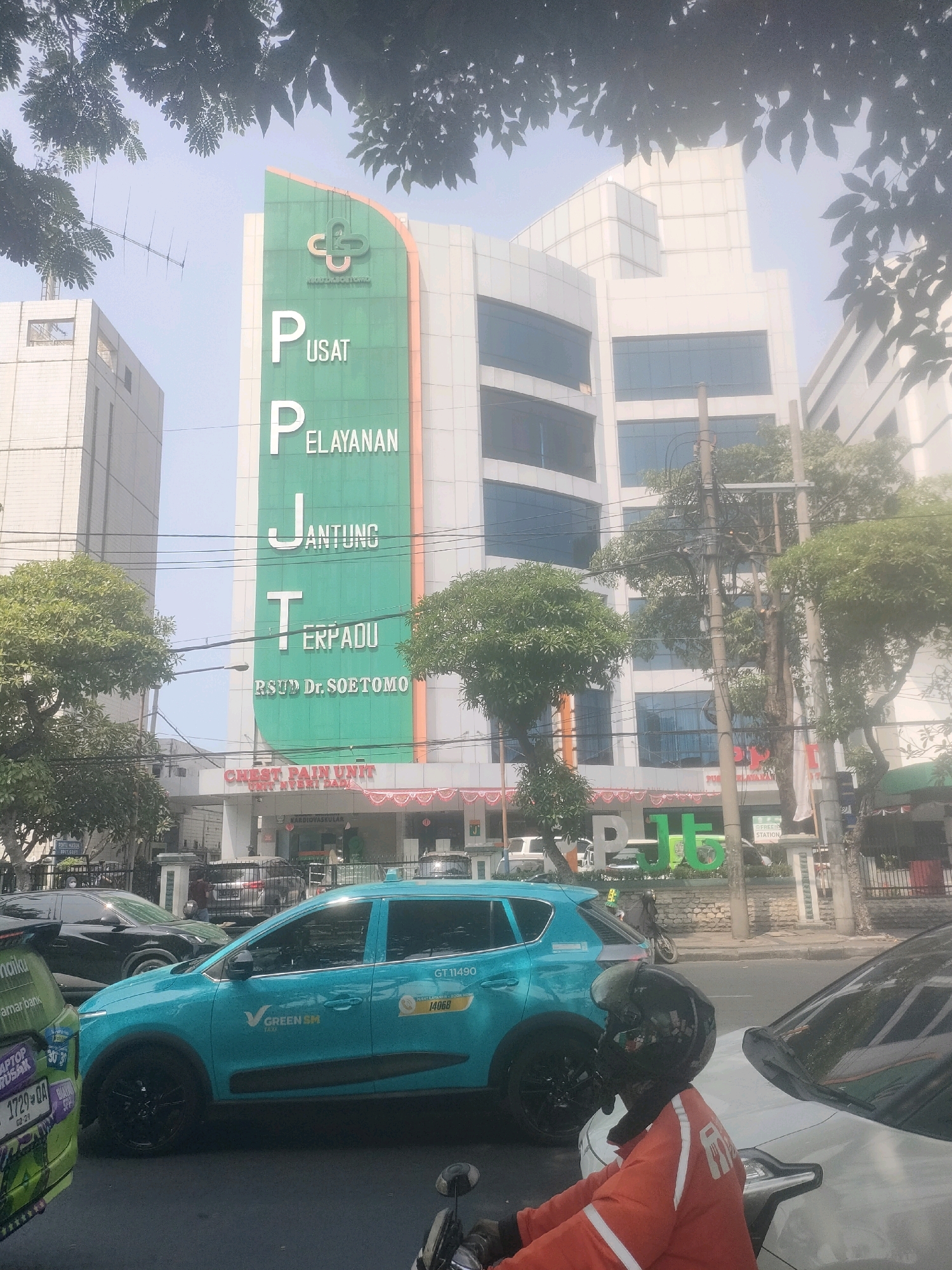 This is a street-level, medium-distance, eye-level shot of a modern hospital building in Surabaya, Indonesia, during daylight. The building prominently displays large, vertical green signage with white text that reads "PUSAT PELAYANAN JANTUNG TERPADU" and below it, "RSUD Dr. SOETOMO". Above this text is a green logo with a stylized heart shape. The building itself is contemporary, with multiple floors and large glass windows.
In the foreground, a teal-colored taxi with "GREEN SM TAXI" branding is visible, stopped in traffic. Behind it, a black car is also stopped. Further back, another green vehicle with various stickers is partially visible. A motorcyclist wearing an orange jacket and a black helmet is positioned in the lower right corner, seemingly waiting in traffic. The environment is urban, with traffic lights, power lines, and trees lining the street. The sky is bright and hazy, suggesting a sunny but potentially humid day. The overall scene depicts a busy urban intersection in front of a major medical facility.