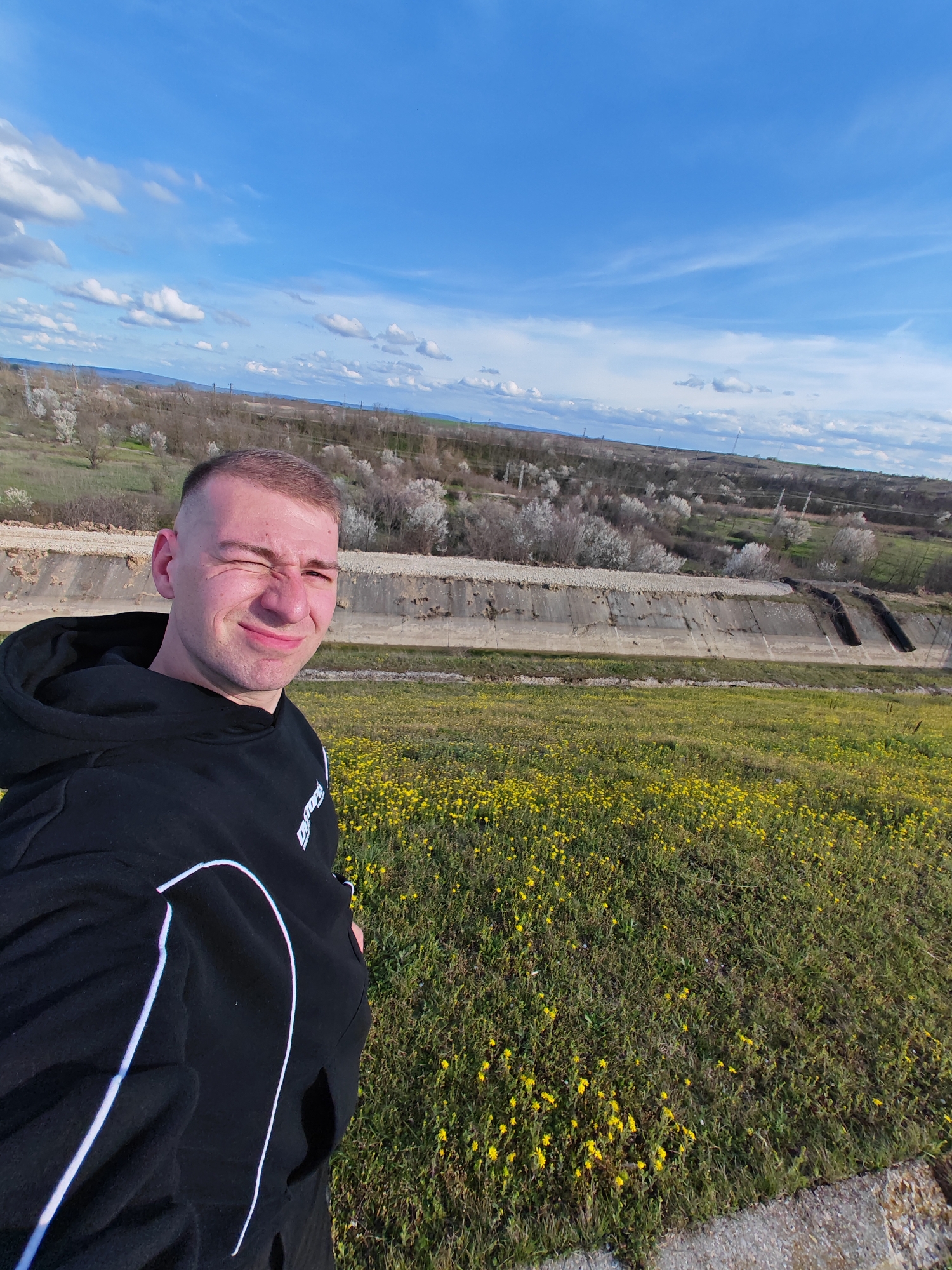 A man is posing for a selfie outdoors. He is fair-skinned with short, cropped hair. He is wearing a black hooded sweatshirt with white stripes. He is winking with his left eye.
The background shows a landscape with a concrete drainage channel. Beyond the channel is vegetation with trees that appear to be in bloom. In the distance are fields and hills under a partly cloudy blue sky.
The location is Shumen, Bulgaria.