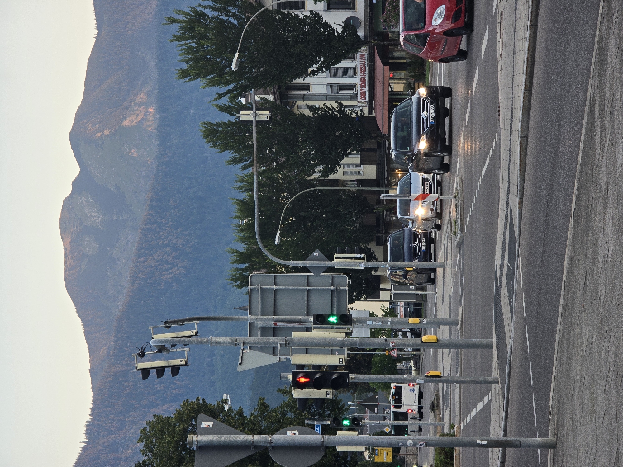 The image shows a street intersection in Garmisch-Partenkirchen, Germany, with traffic lights and several cars. In the foreground, a red car is stopped at a red light, while a black SUV and a silver sedan are also present in the intersection. Traffic lights are visible, with one showing a red light and a pedestrian figure, and another showing a green light with a walking figure. A white bus is visible in the background. The scene is set against a backdrop of trees and mountains, suggesting a location near the Alps. The sky is overcast, indicating it is likely late afternoon or early evening. The road appears to be wet, reflecting the car headlights and traffic lights.